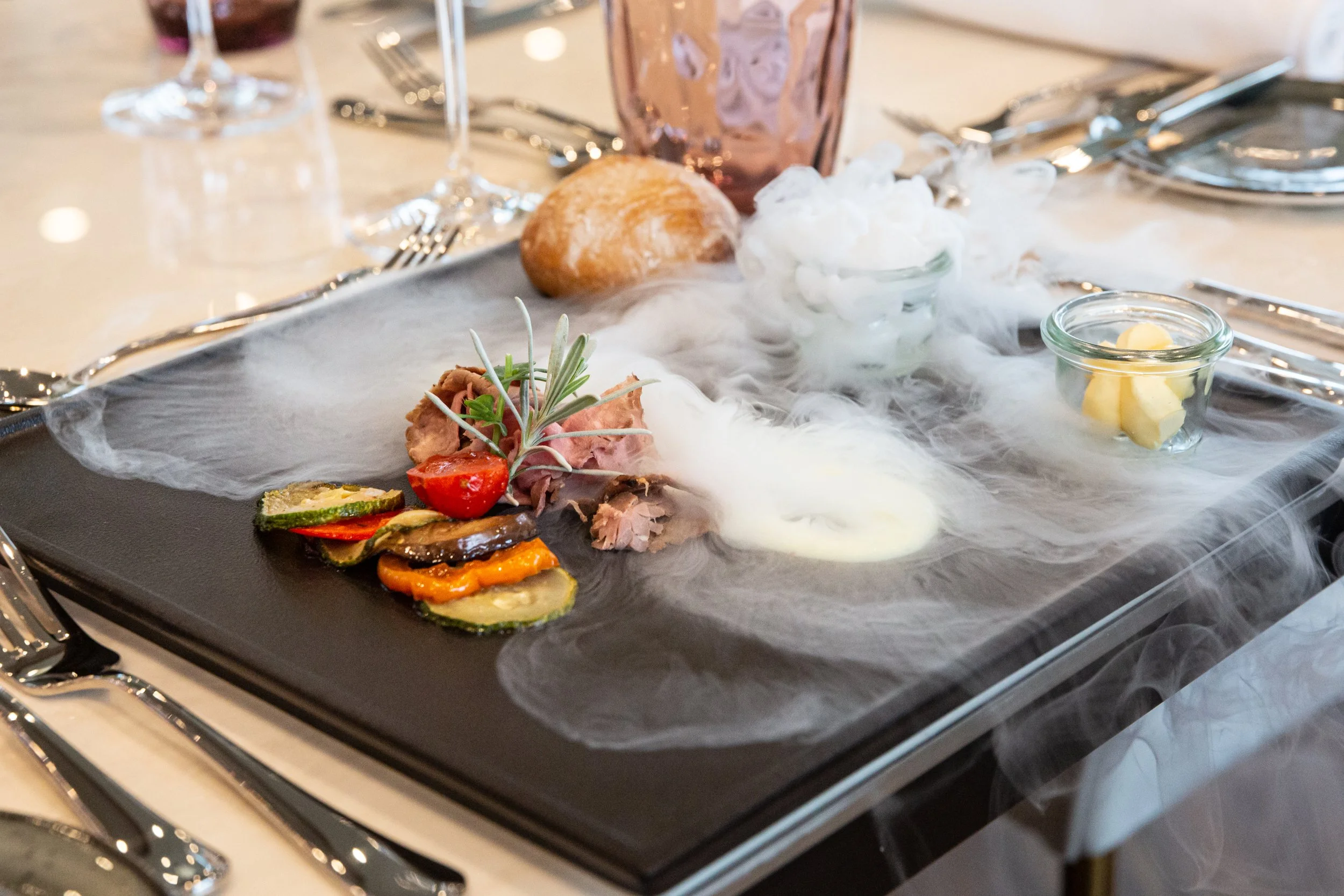 Fein angerichteter Gourmetteller mit rosa gebratenem Fleisch, gegrilltem Gemüse und Rosmarinzweig, umgeben von dramatischem Nebel-Effekt auf schwarzem Teller