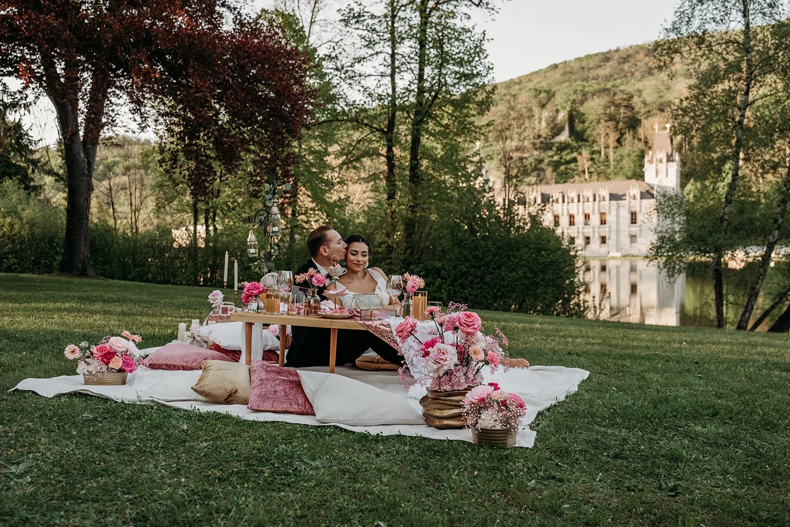 Romantischer Heiratsantrag im Park von Schloss Hernstein: Ein Paar sitzt auf Kissen an einem niedrigen Tisch, umgeben von rosa Blumenarrangements, Kerzen und dekorierten Gläsern, während im Hintergrund Bäume und ein Gebäude zu sehen sind