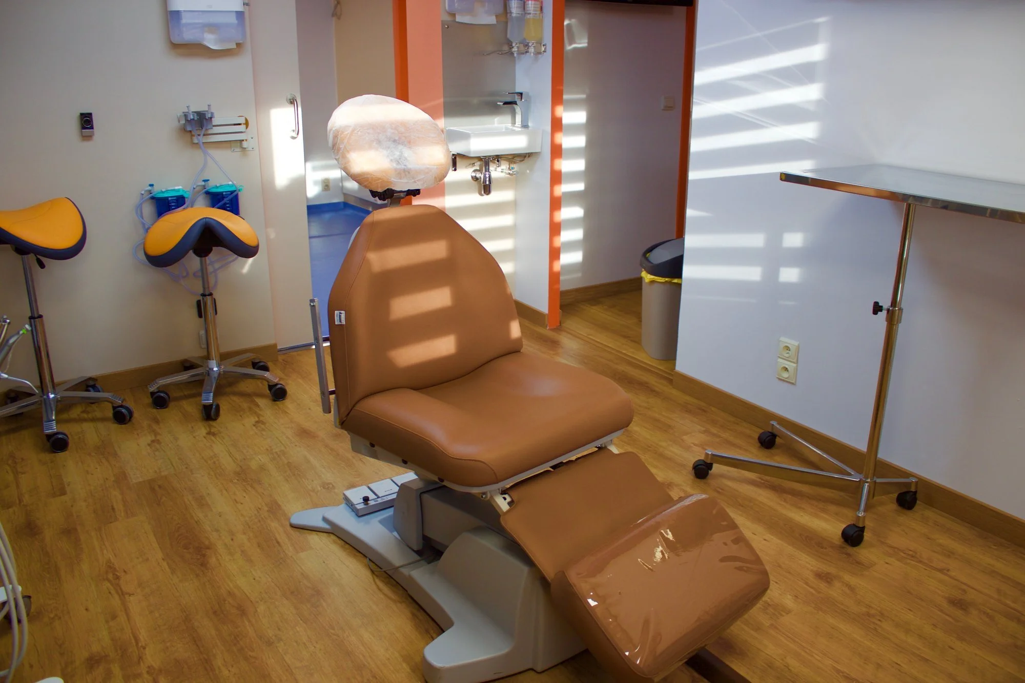 Empty dental examination room with a brown dental chair, yellow and blue dental stools, a small sink, trash can, and medical equipment, in a well-lit, clean setting.
