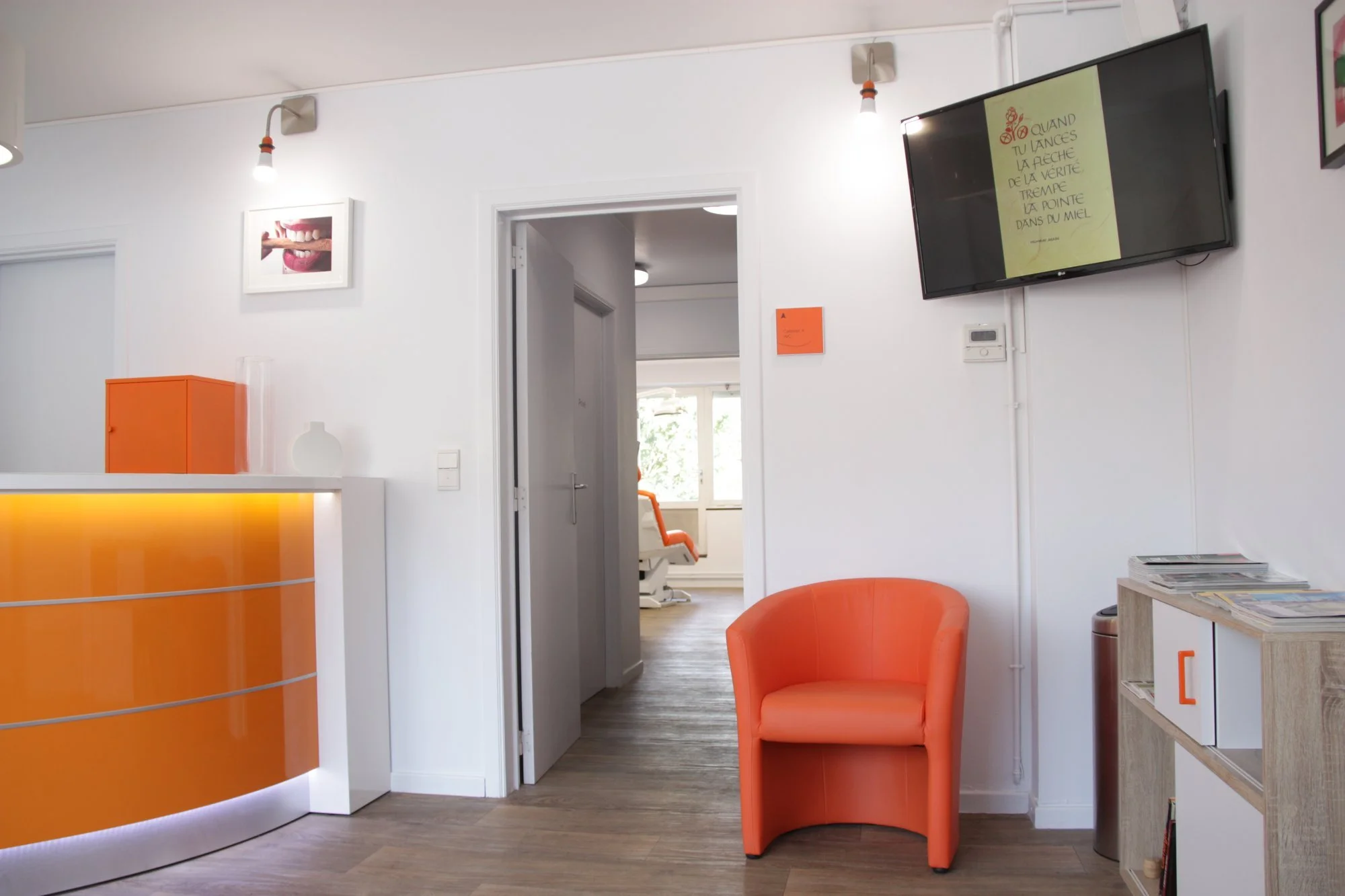 Interior of a dental clinic or medical office with a bright reception area. Features include an orange and white front desk, an orange chair, wall-mounted TV displaying text in French, dental chair visible through an open door, artwork on white walls