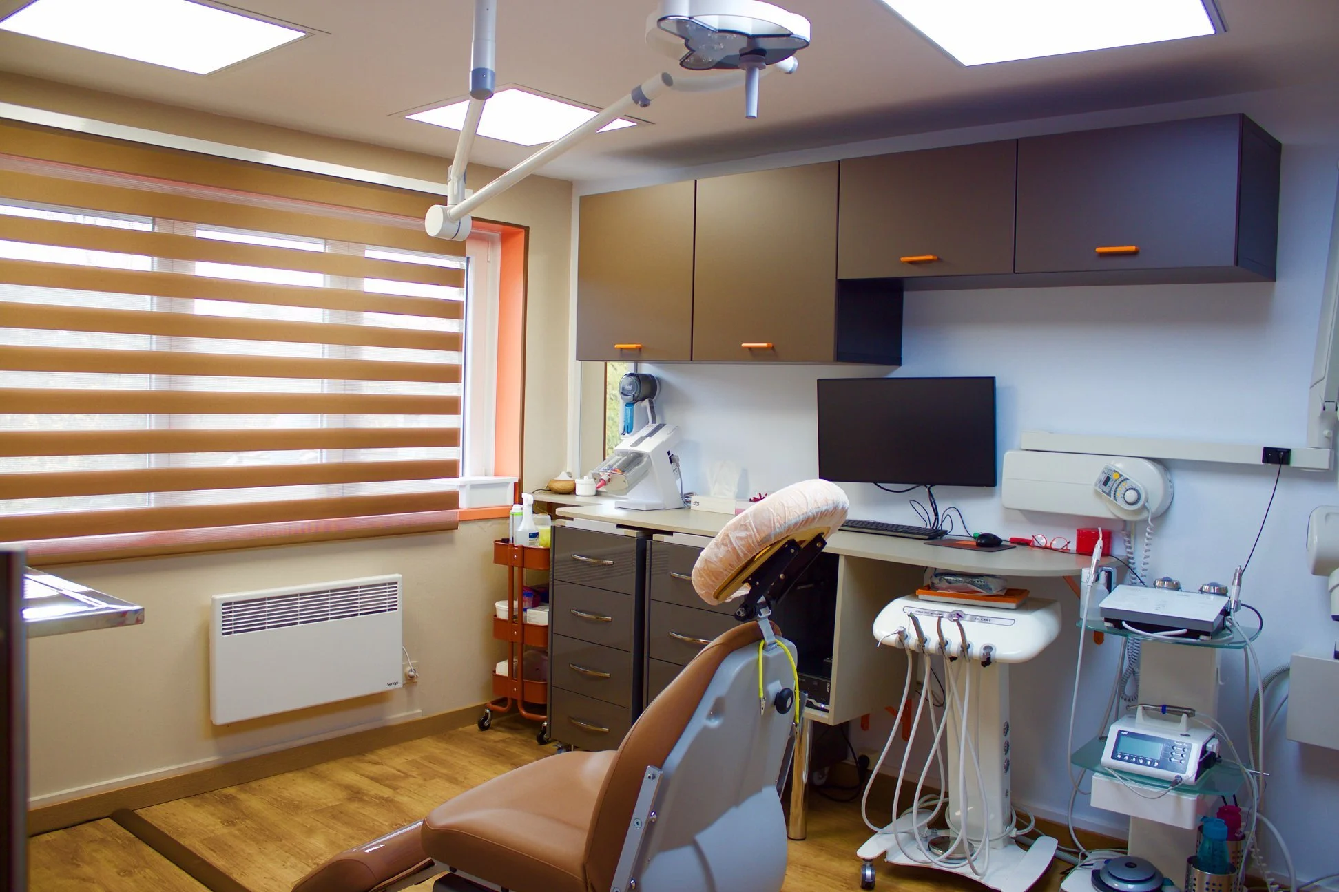 Dental office with dental chair, monitor, overhead light, storage cabinet, medical equipment, and window with blinds.