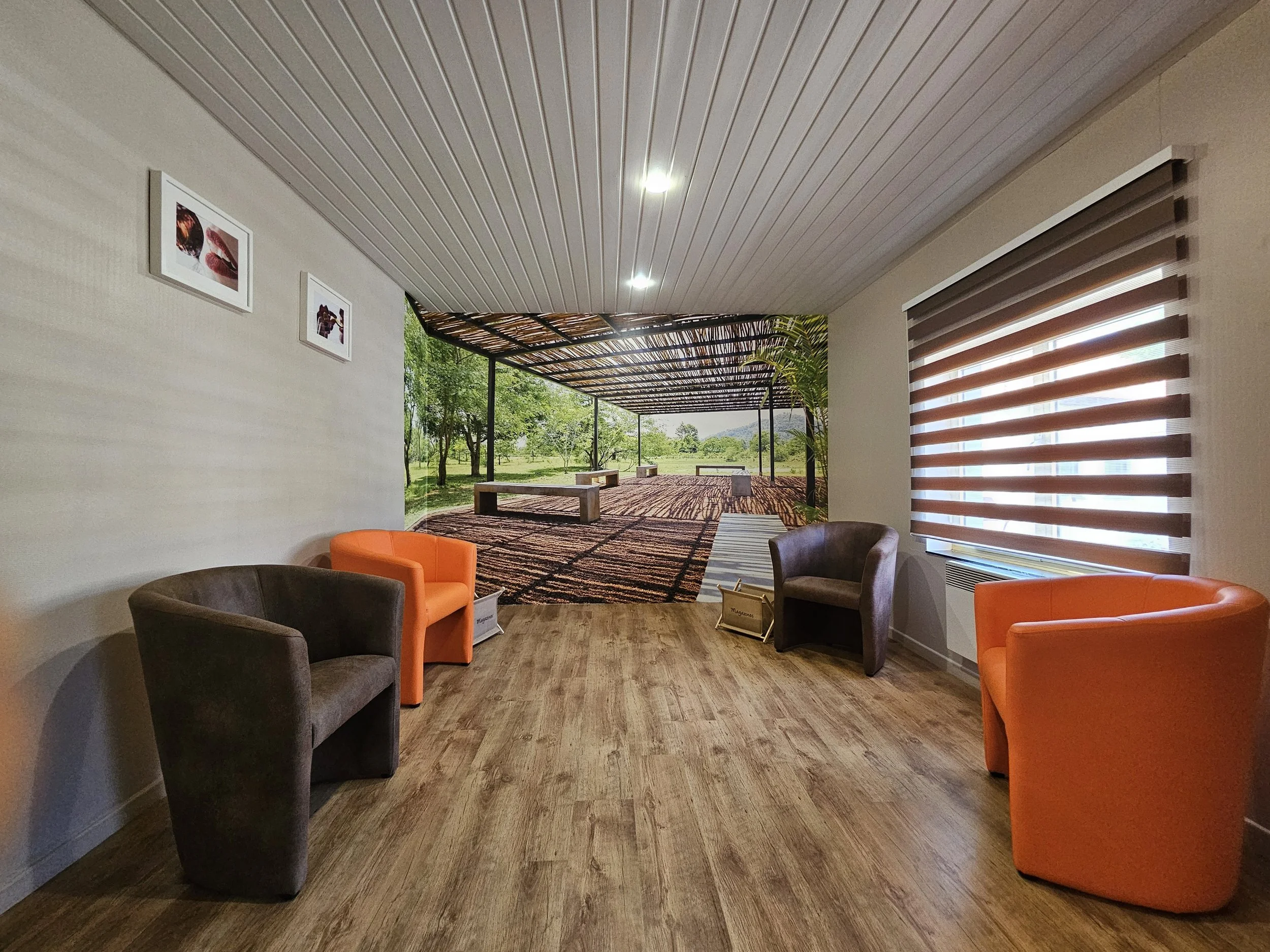 A cozy room with four chairs (two brown and two orange), two small tables, a wall-mounted art with three pictures, and a large window with brown blinds. A large outdoor mural depicts a patio with benches, trees, and a roof, creating an indoor-outdoor