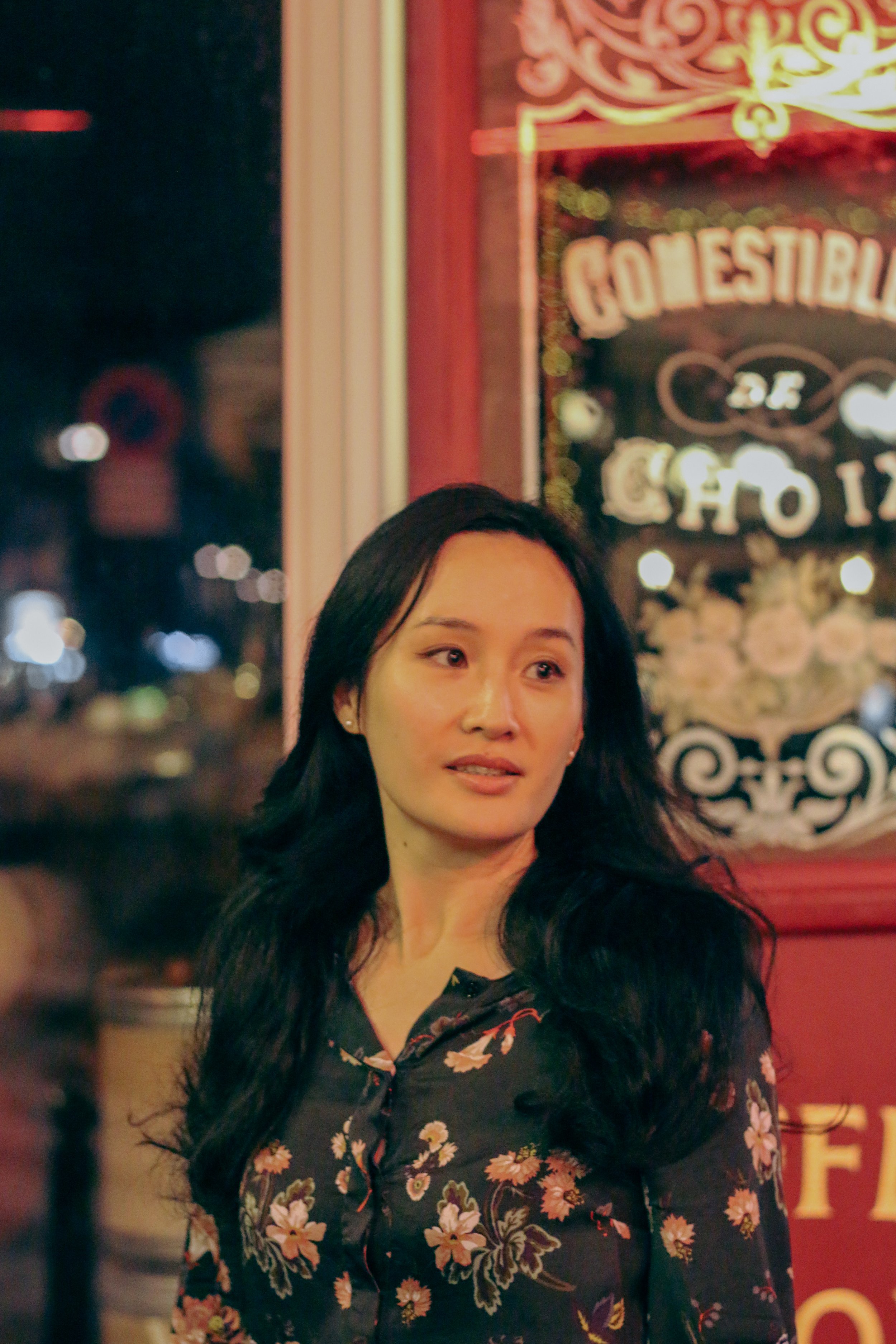 Une femme aux cheveux longs et noirs, portant une chemise à fleurs, se tient dans un lieu lumineux avec un décor coloré en arrière-plan, notamment un tableau ou une pancarte avec le mot 'GÓMEStIBIL'.
