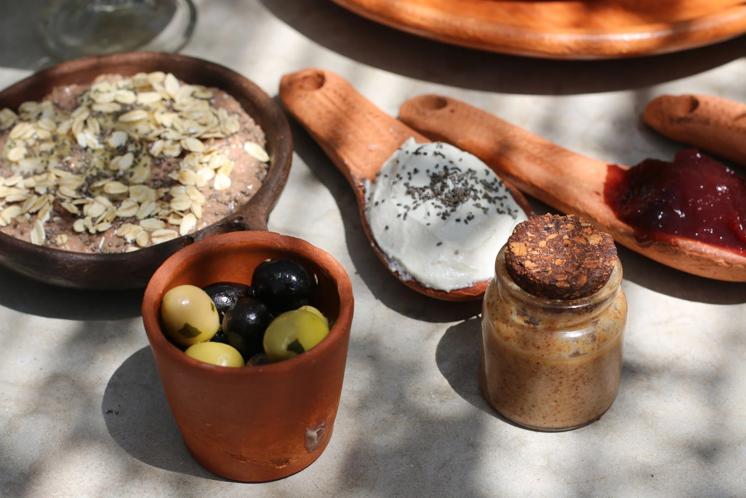 Différents aliments traditionnels grecs sur une surface en pierre, notamment du yaourt nature avec des graines de chia, un petit pot de fromage, des olives vertes et noires, et diverses tartines avec confiture ou beurre.