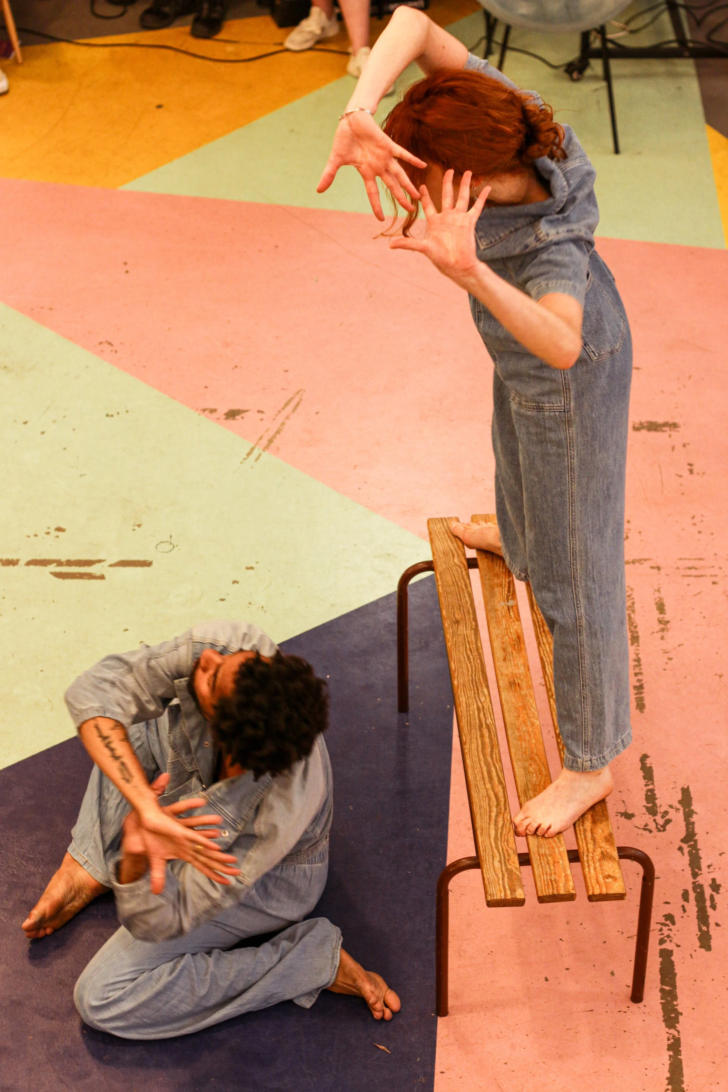 Deux personnes en costume de scène, l'une debout sur un banc en bois, l'autre assise au sol, en train de jouer une scène dramatique ou comique dans un espace coloré.