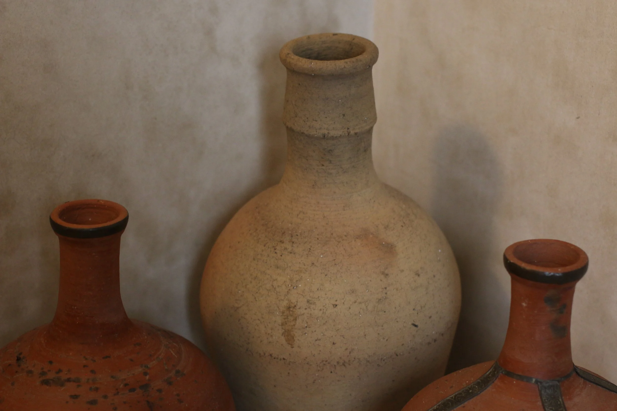 Trois anciennes jarres en terre cuite, deux de couleur rouge et une de couleur beige, disposées contre un mur beige.