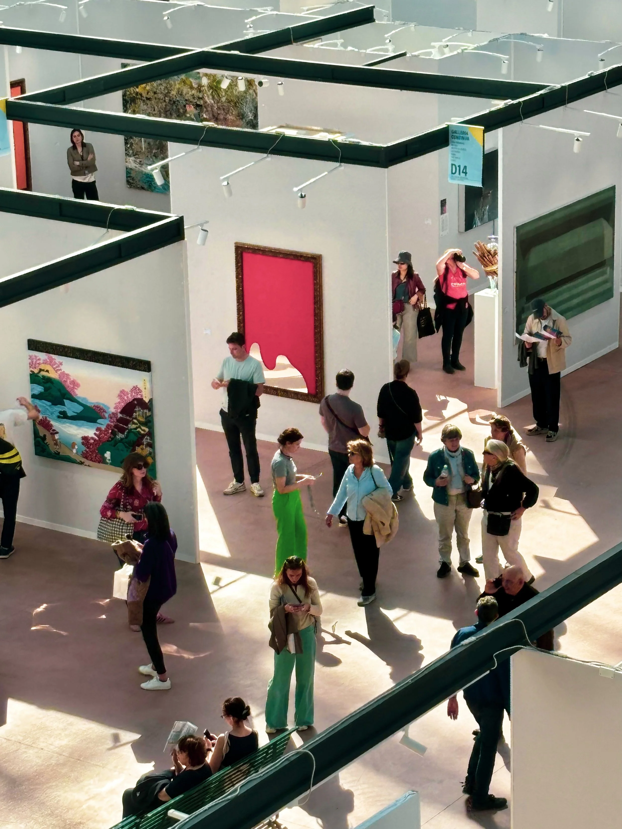 Une galerie d'art avec des visiteurs admirant diverses œuvres, y compris une peinture avec un paysage de montagne et une toile rose encadrée. Des gens discutent, lisent ou prennent des photos dans un espace bien éclairé.