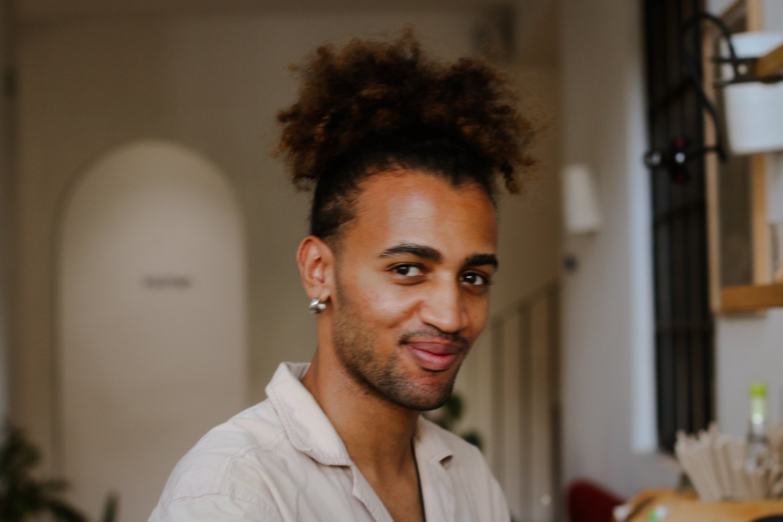 Jeune homme avec des cheveux afro coiffés en haut, portant une chemise beige, dans un intérieur lumineux.