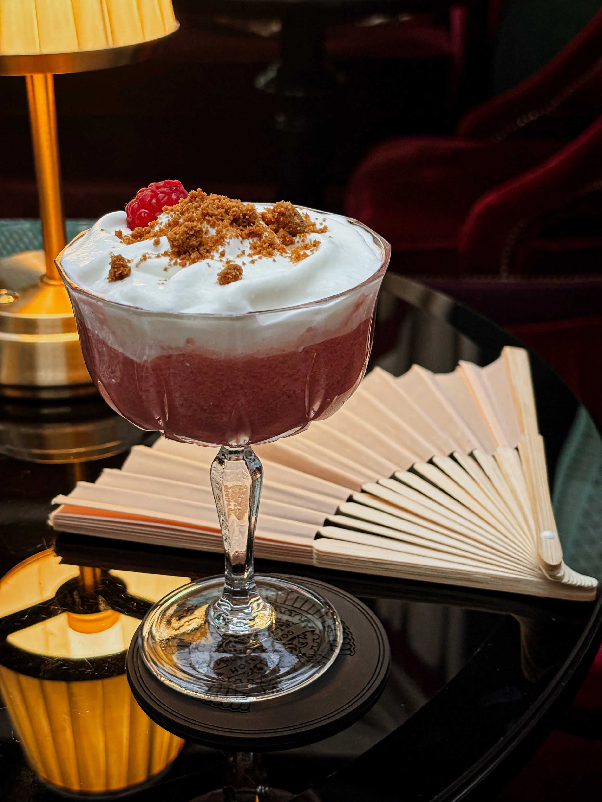 Verre à cocktail rempli d'une boisson rouge avec de la crème fouettée, des miettes et une framboise dessus, posé sur une table avec un livre ouvert en arrière-plan, éclairé par une lampe