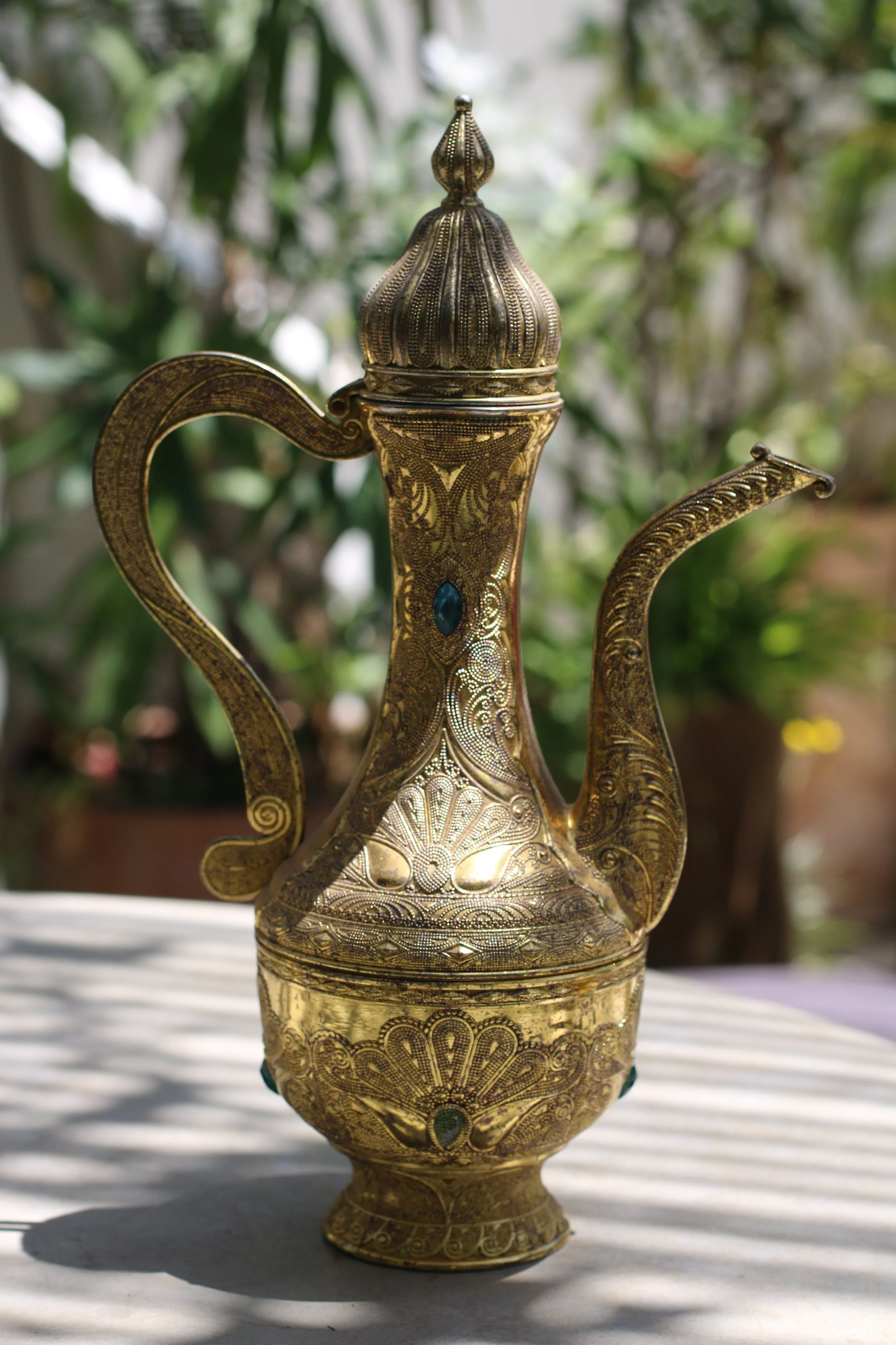 Une théière en métal doré avec des motifs élaborés et des pierres décoratives, posée sur une surface en plein air avec un fond flou de végétation.