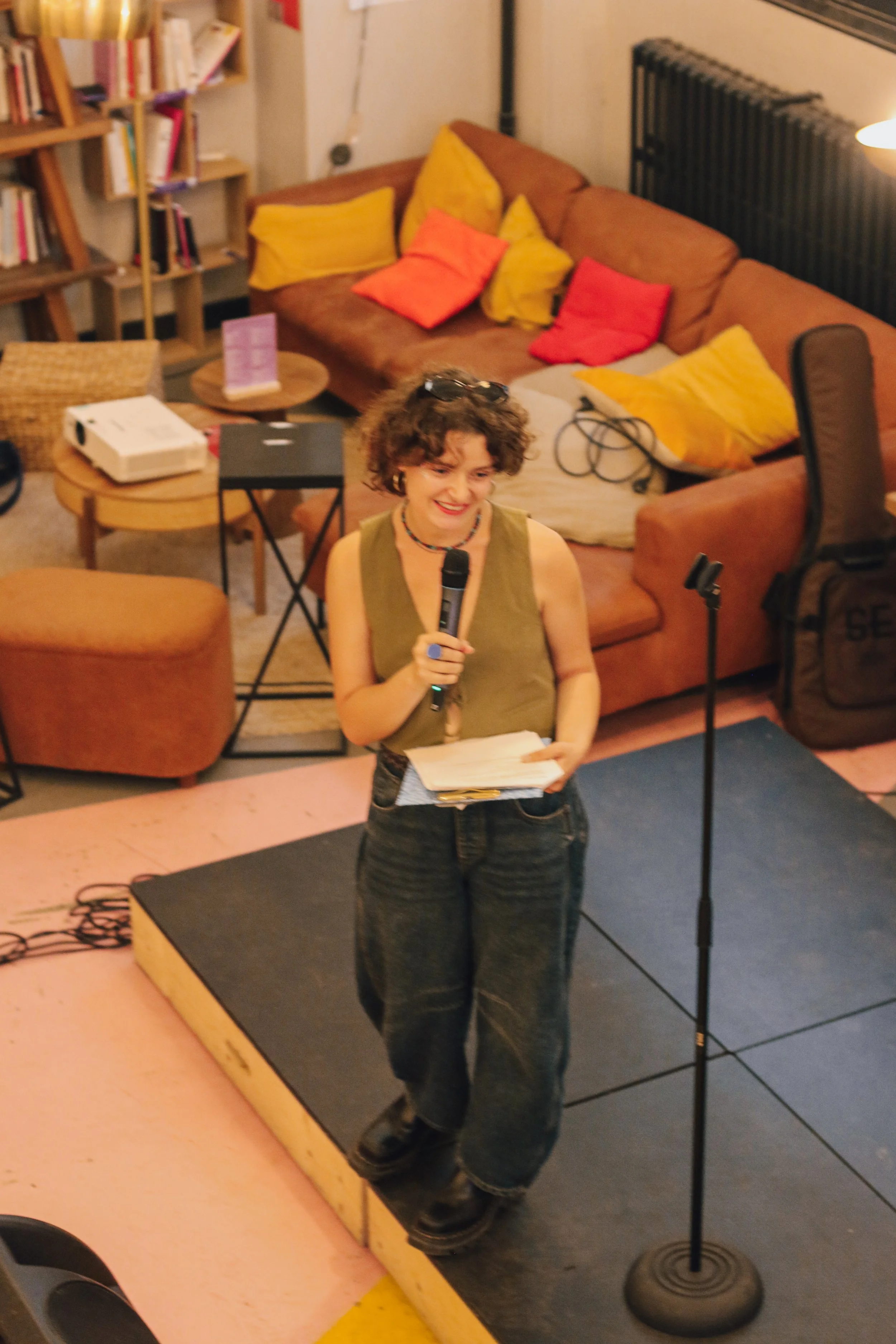 Une femme souriante tenant un micro et des notes, se tenant sur une petite scène dans un salon accueillant avec un canapé en laine orange et des coussins colorés, des étagères remplies de livres en arrière-plan.
