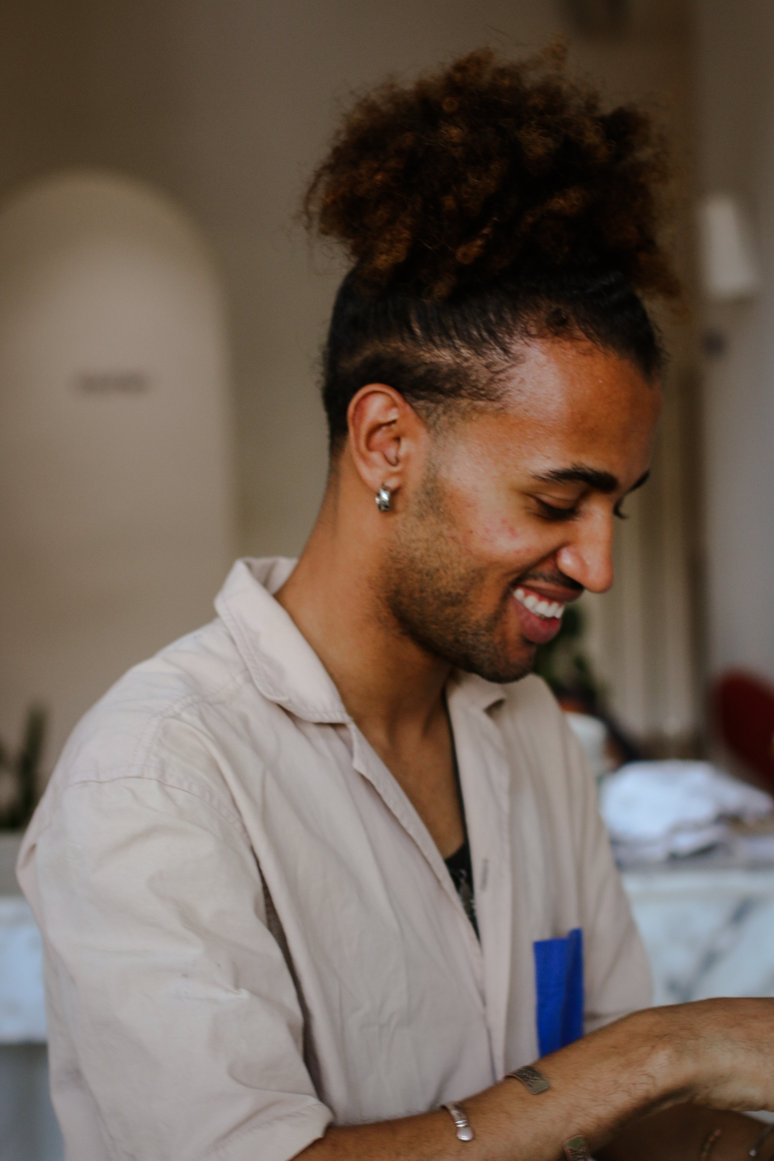 Un homme souriant avec des cheveux en chignon et portant une chemise beige, intérieur chaleureux.