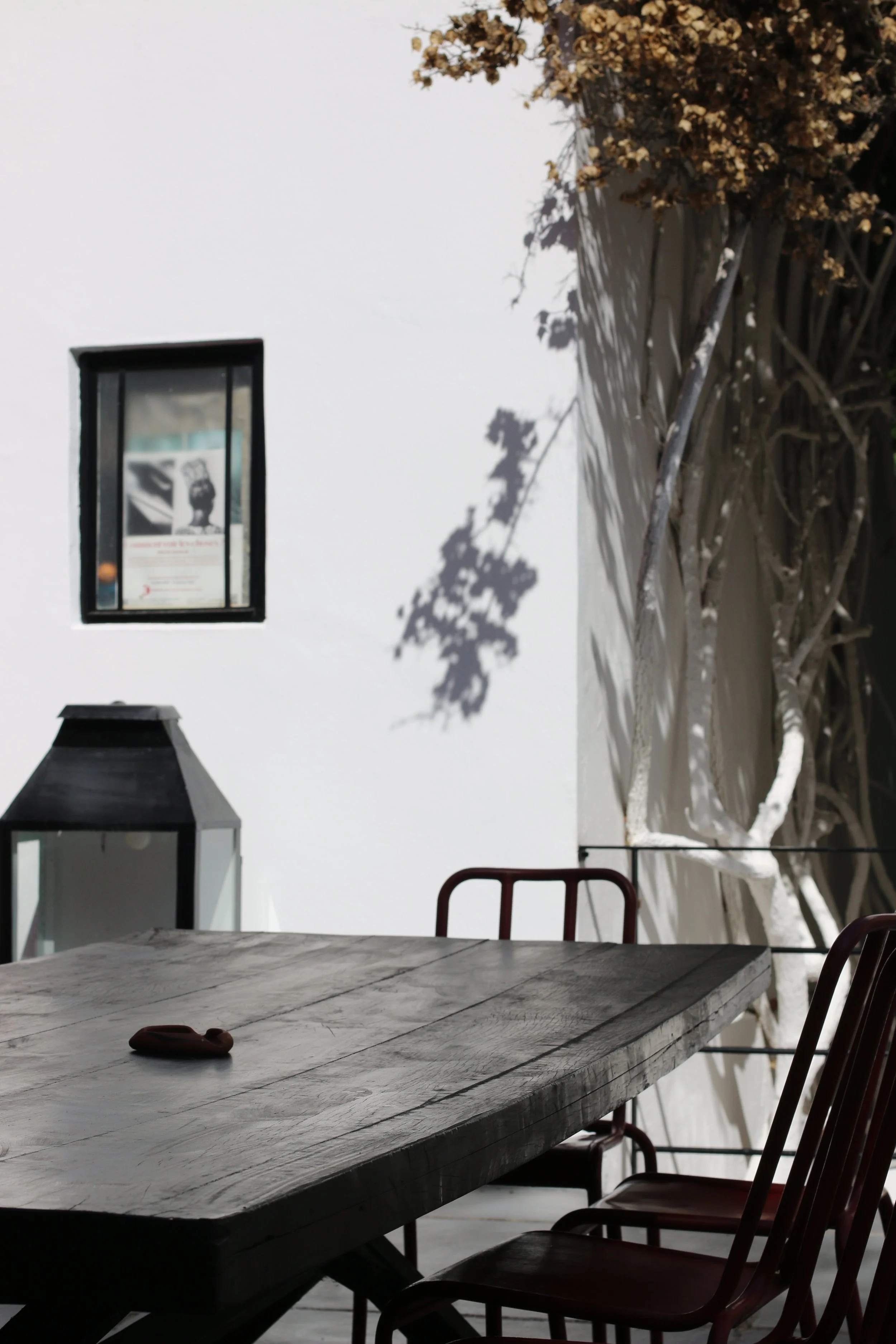 Une table en bois avec quatre chaises en métal, une lanterne noire et une fenêtre dans un mur blanc, avec des branches d'arbres projetant des ombres sur le mur.