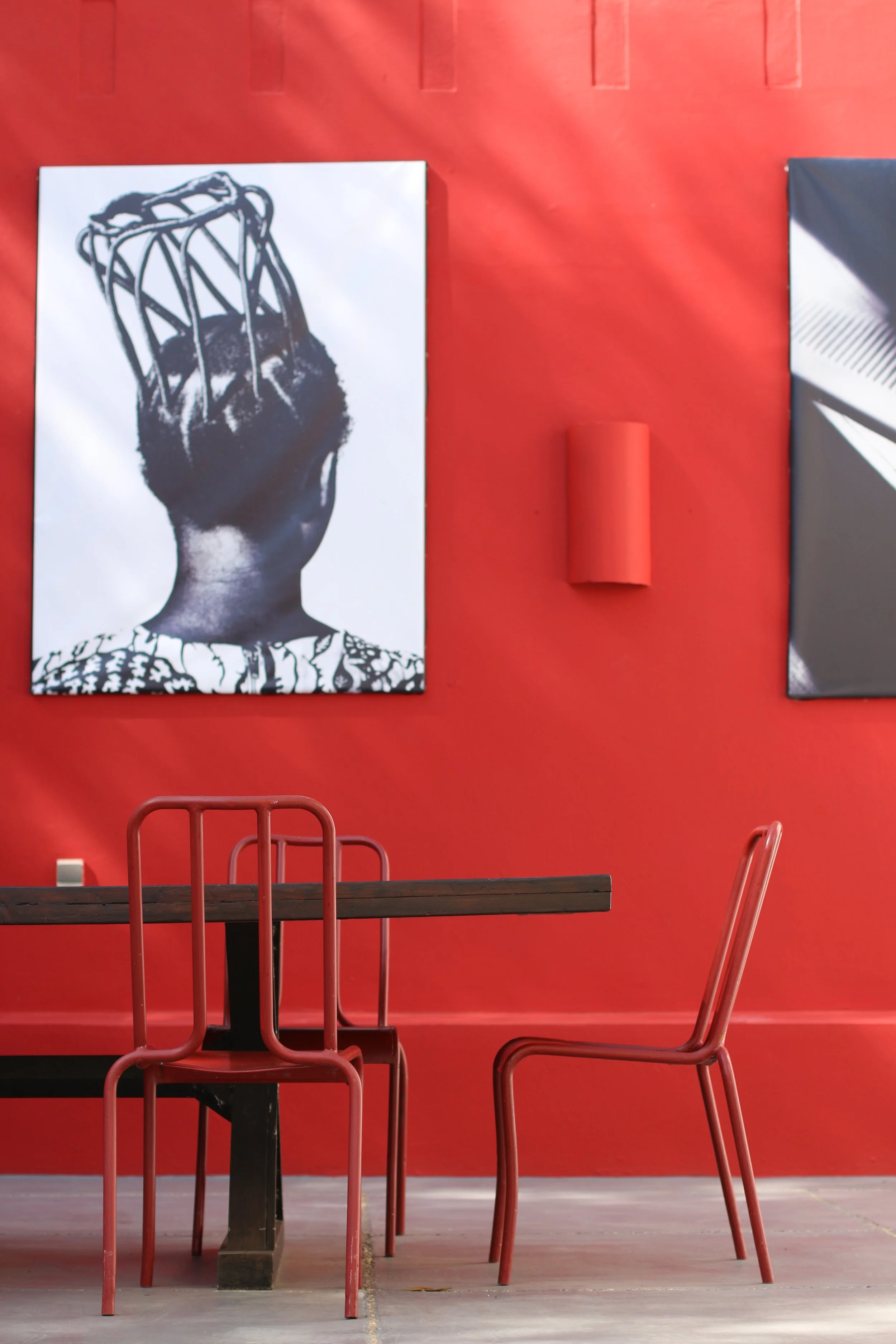 Une salle avec une table en bois et deux chaises en métal rouge, mur rouge avec deux œuvres d'art modernes, une grande photo en noir et blanc d'une personne avec des tresses en forme de cage. La lumière naturelle crée des reflets et des motifs d'ombr