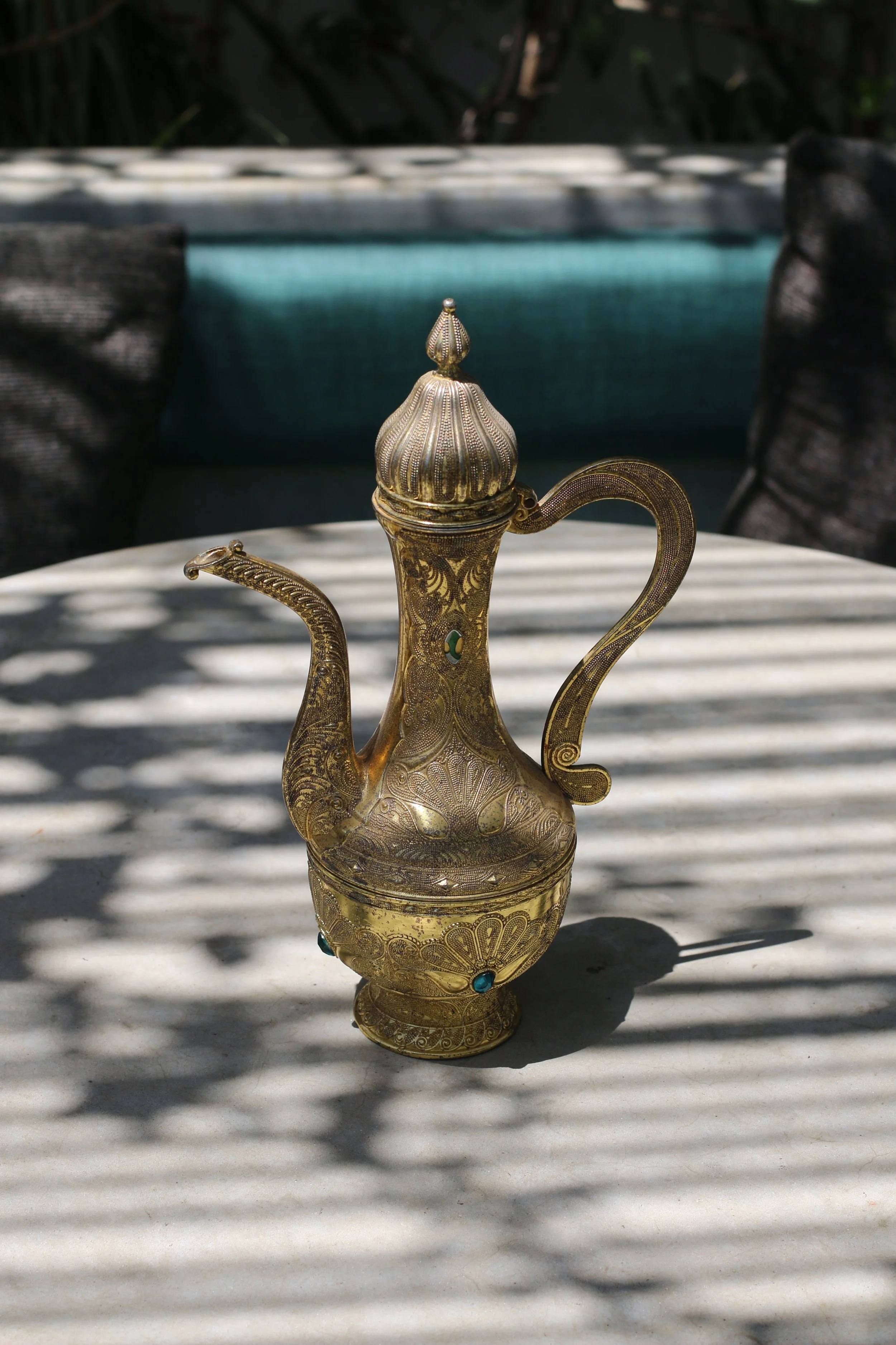 Une théière en métal doré ornée de gravures complexes, posée sur une table en extérieur avec des ombres de feuillage en motif sur la surface.