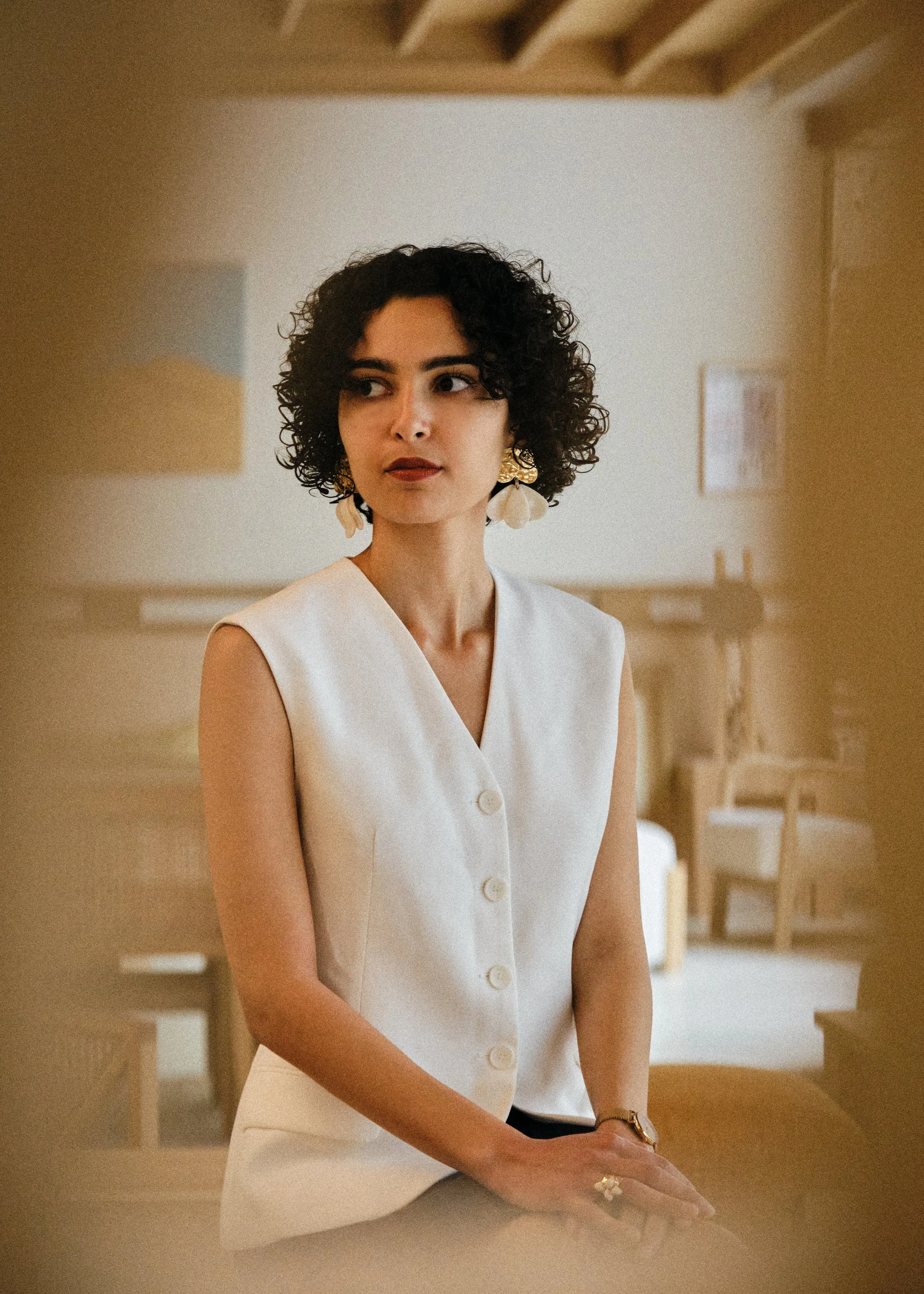 Une femme aux cheveux bouclés, portant une veste blanche à boutons, avec de grosses boucles d'oreilles, se tient dans une pièce chaleureuse avec un plafond en bois.