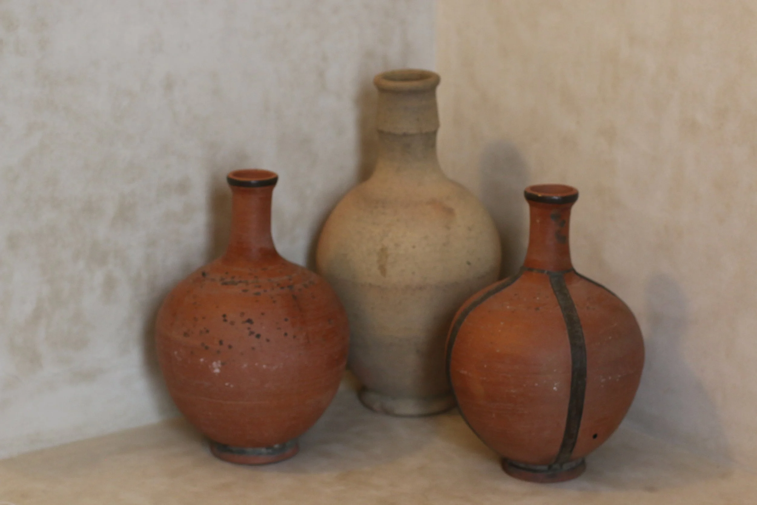 Trois anciennes amphores en terre cuite de différentes tailles et couleurs, exposées sur une surface contre un mur beige.