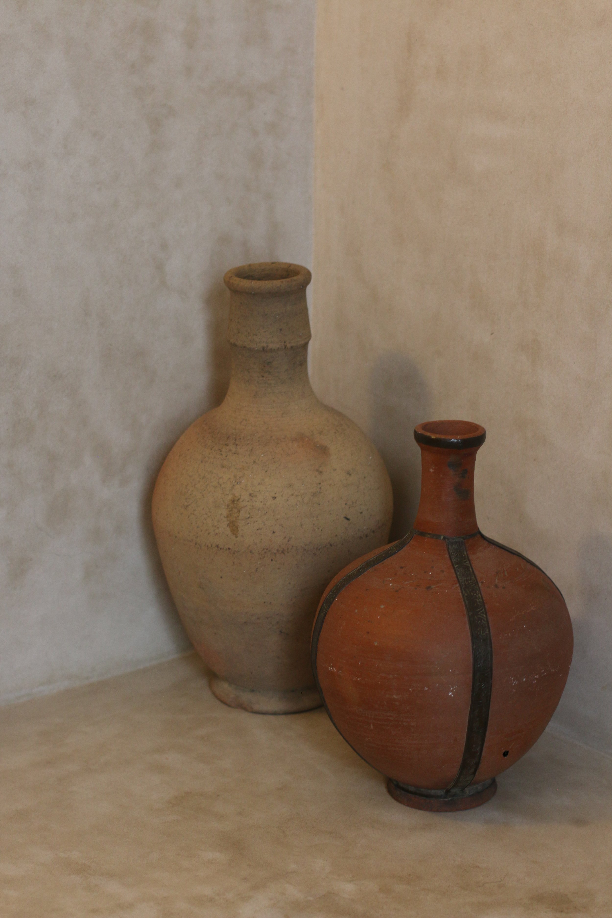 Deux vases en céramique, un beige et un rouge foncé, placés dans un coin contre un mur beige clair.