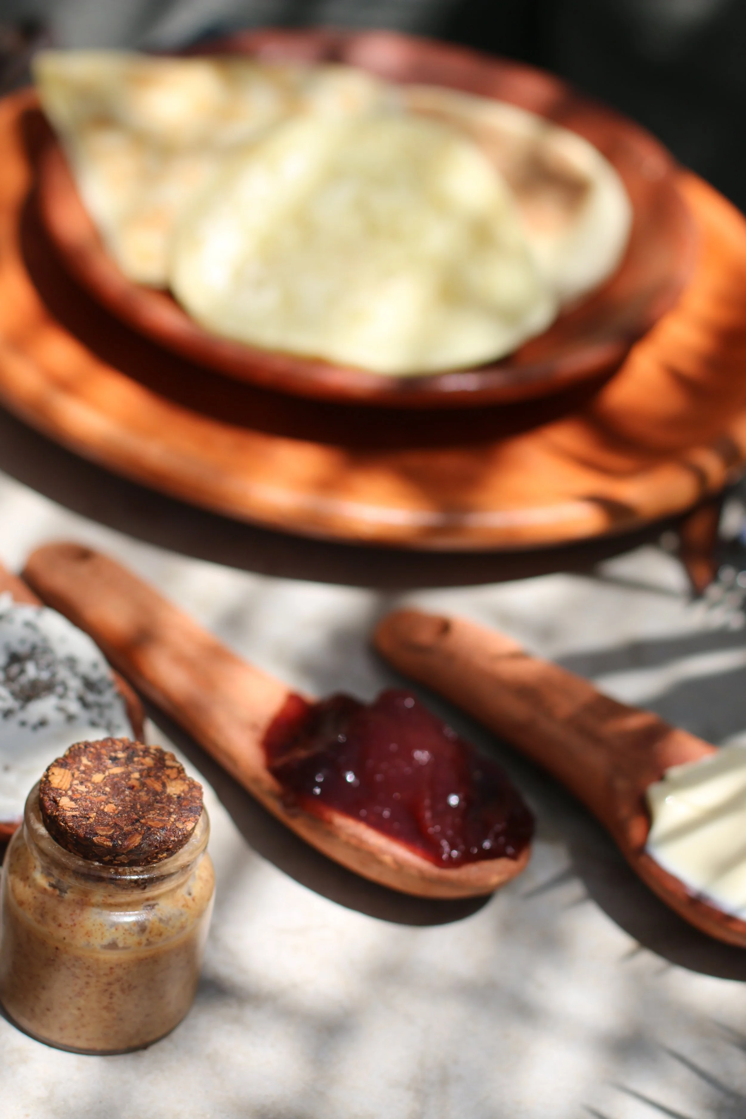 Différents types de confitures et garnitures dans des cuillères en bois, avec un pot de miel ou de beurre et des biscuits en arrière-plan sur une surface claire.