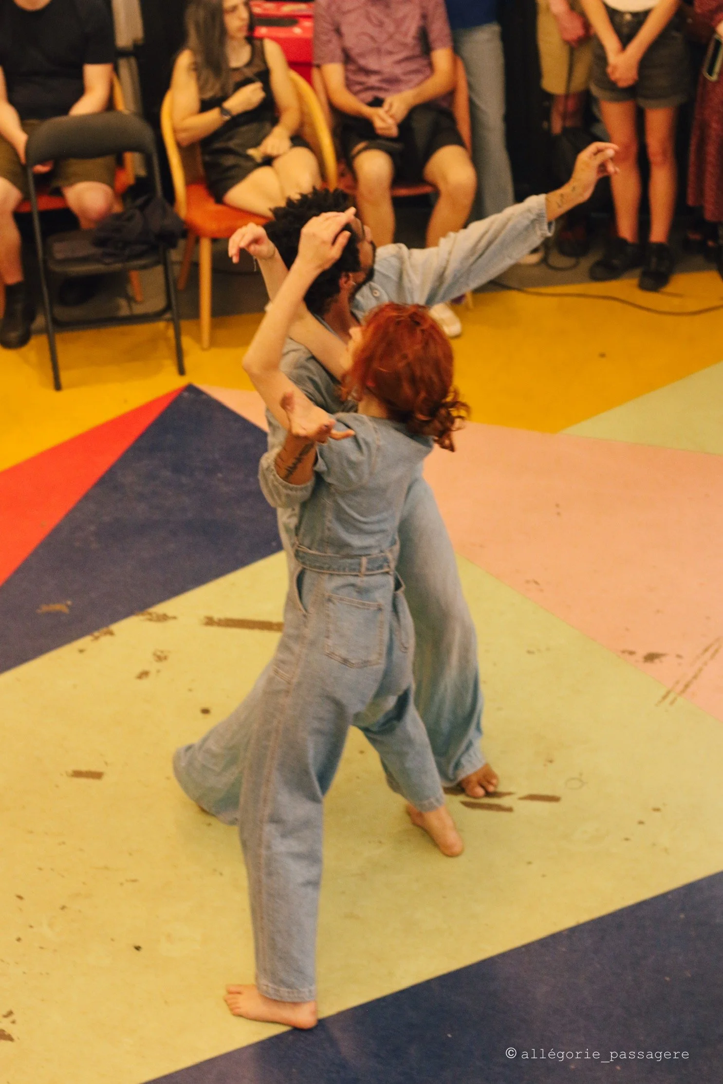 Deux personnes dansantes en denim, entourées de spectateurs assis et debout, salle colorée.