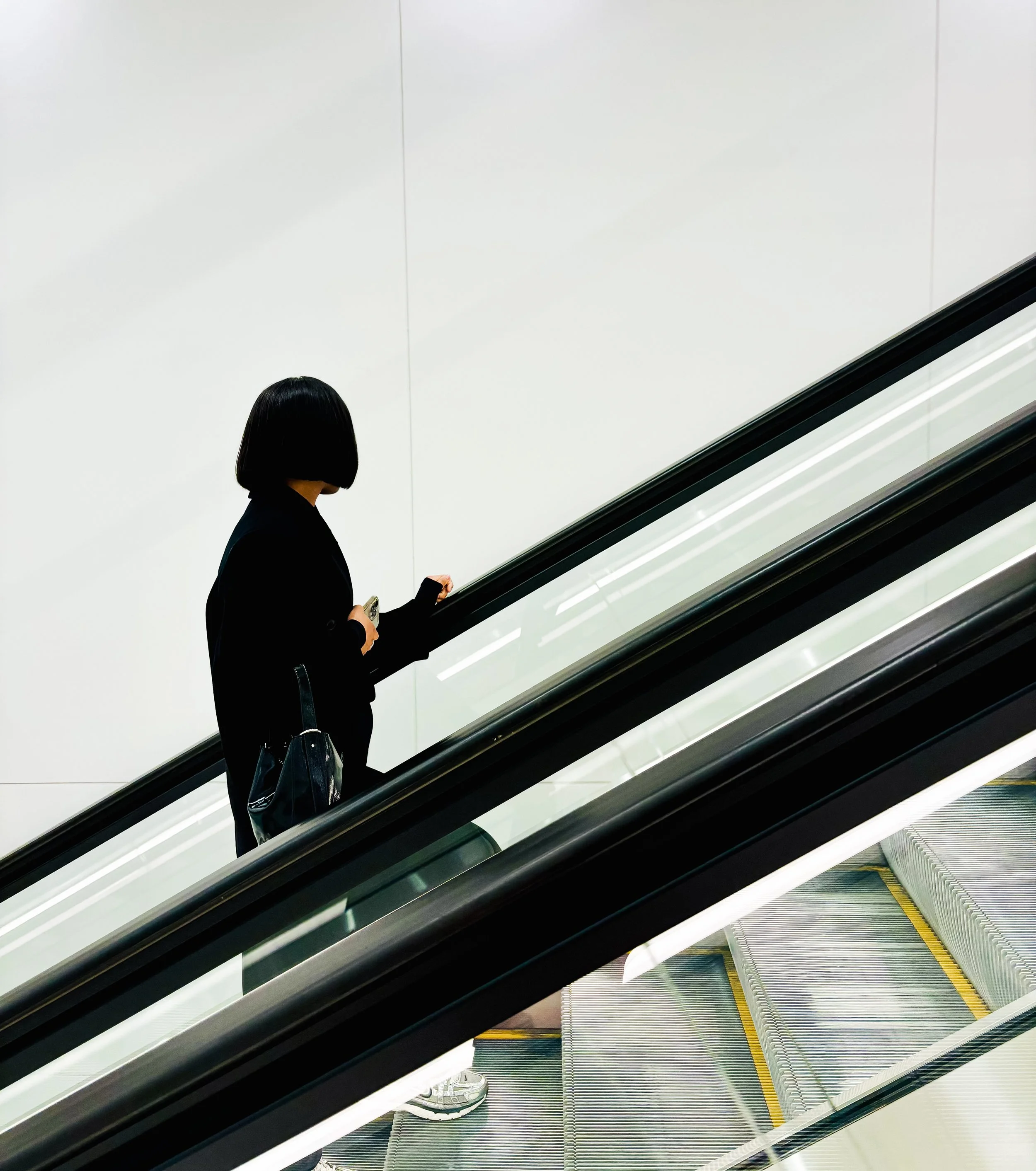 Une femme avec une frange noire sur un escalator, tenant un téléphone portable, portant un manteau noir et une sacoche noire.