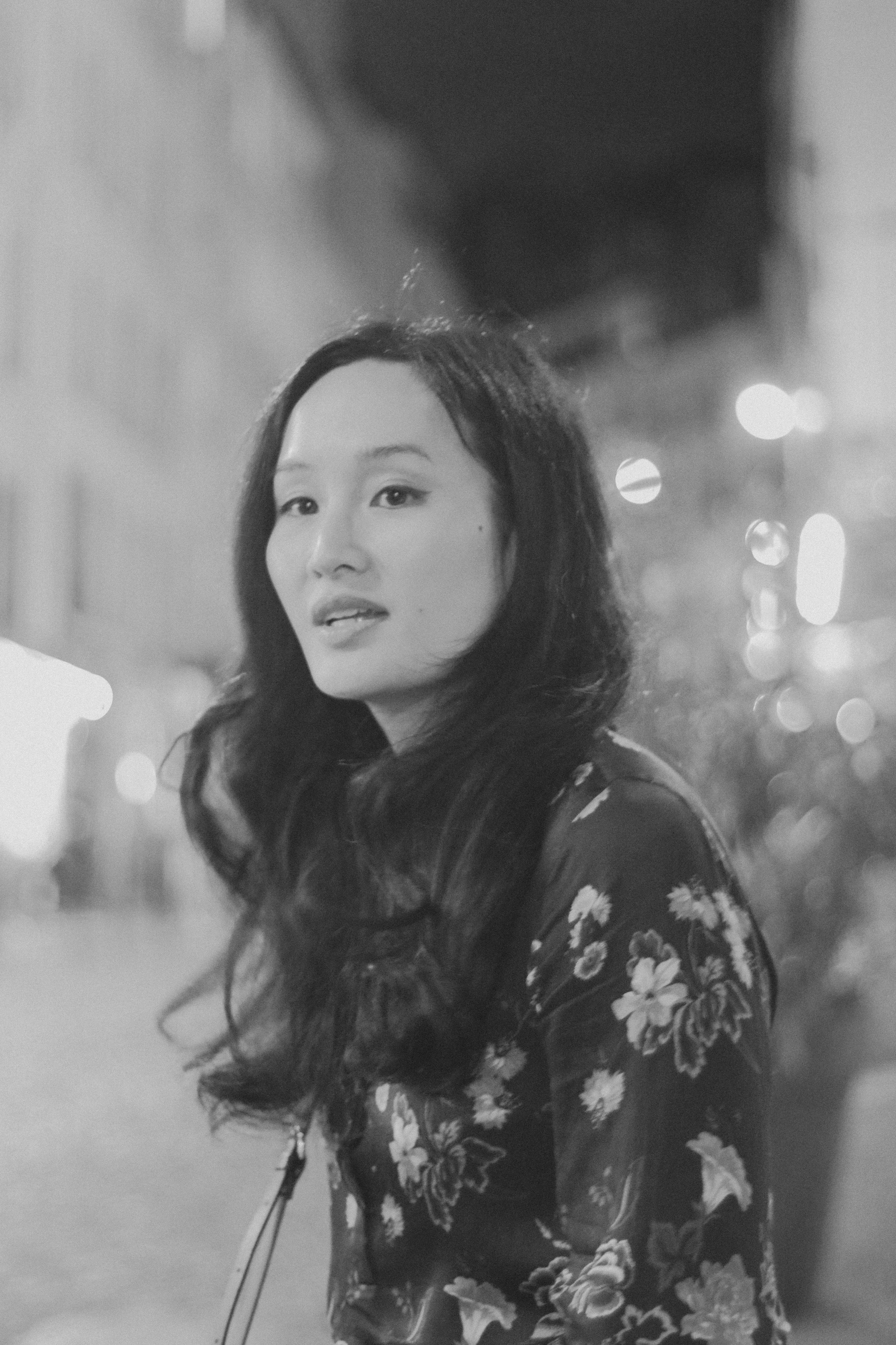 Portrait noir et blanc d'une femme avec de longues cheveux ondulés, portant une robe à motifs floraux, dans une scène urbaine nocturne avec lumières floues en arrière-plan.