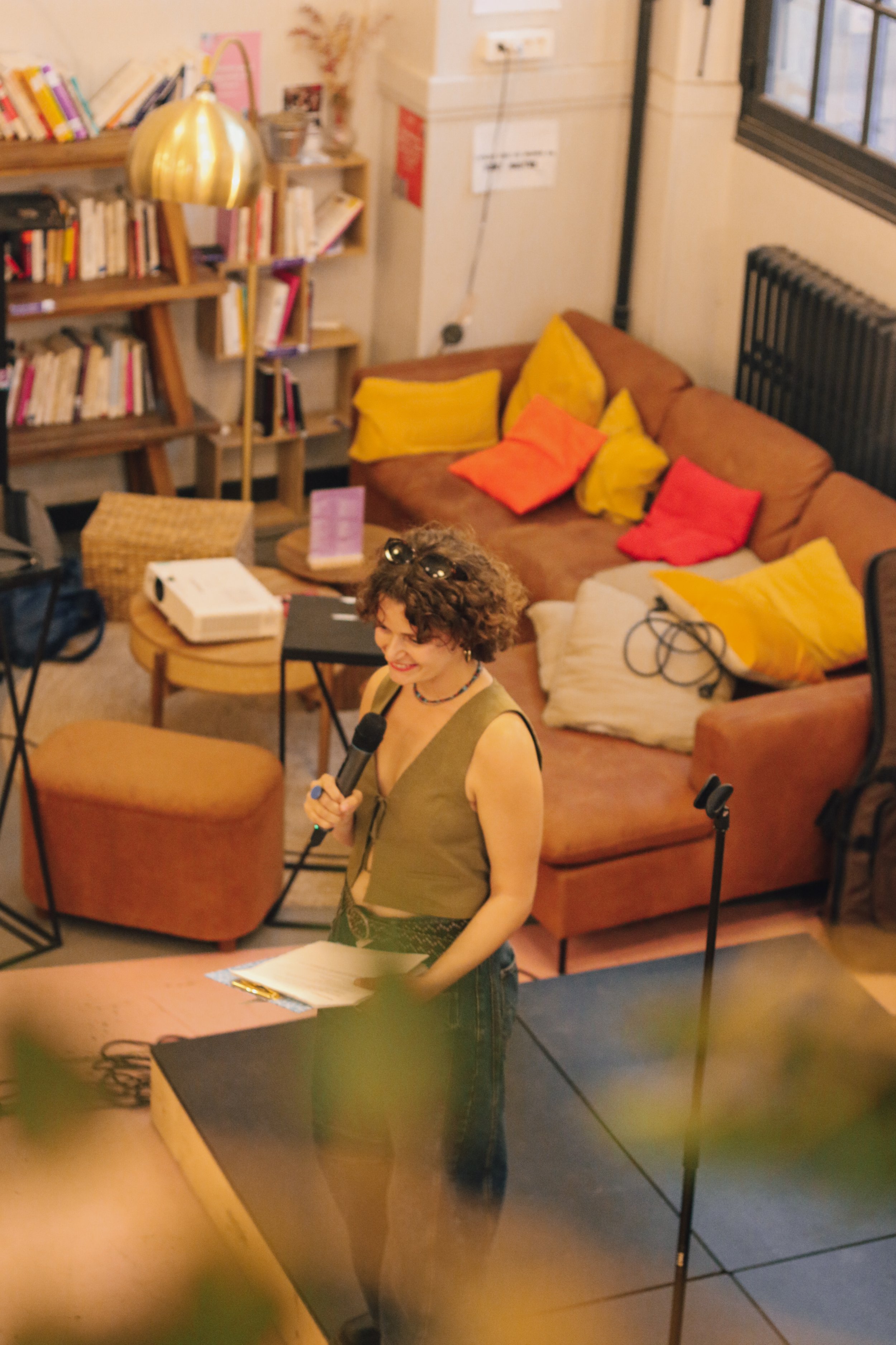 Jeune femme avec micro, parlant ou chantant, dans un salon cosy avec canapé, coussins colorés et étagères remplis de livres.