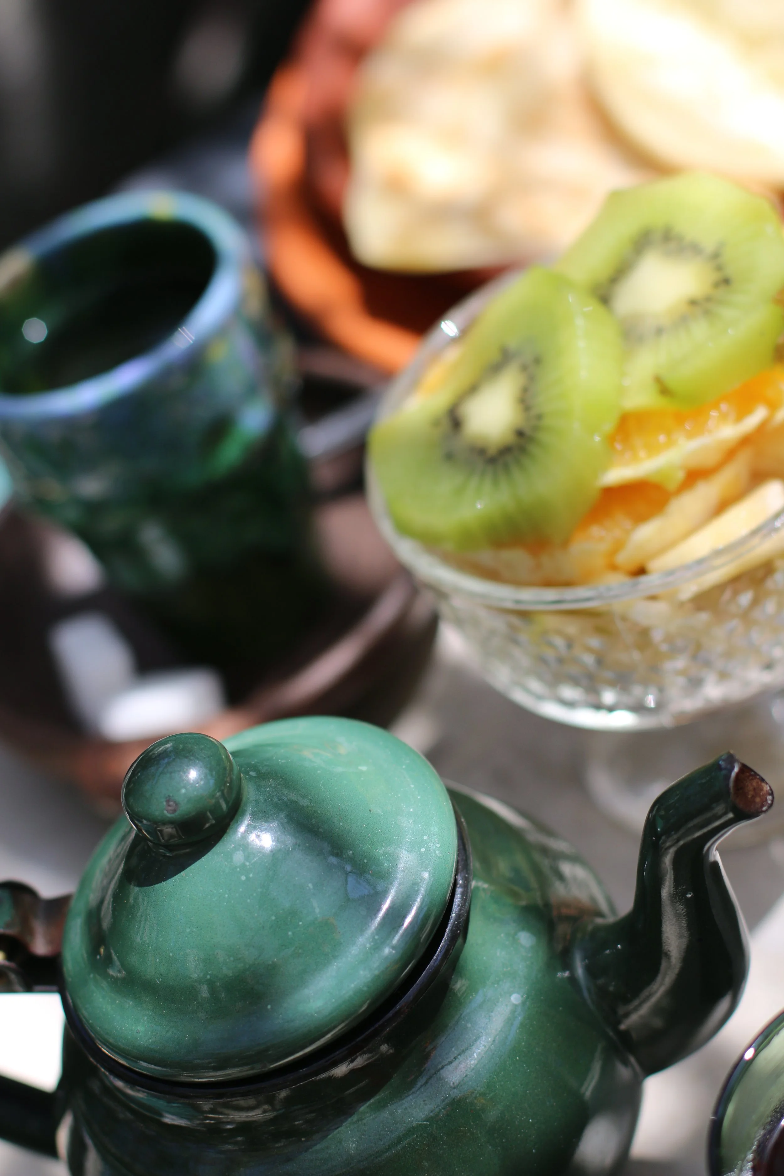 Couvercle vert d'une théière en céramique sur une table avec un bol de fruits comprenant kiwi, mangue et ananas et une tasse en céramique verte