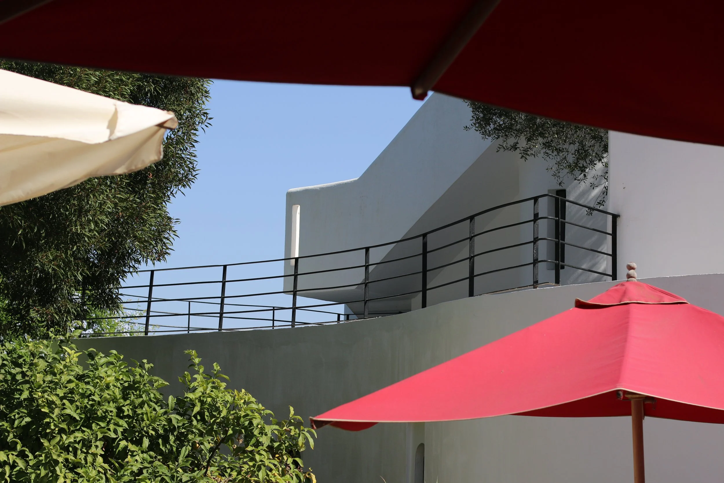Vue partielle de bâtiments blancs avec balustrades en métal, entourés de parasols rouges et beige sous un ciel bleu, avec des arbres verts à l'avant.