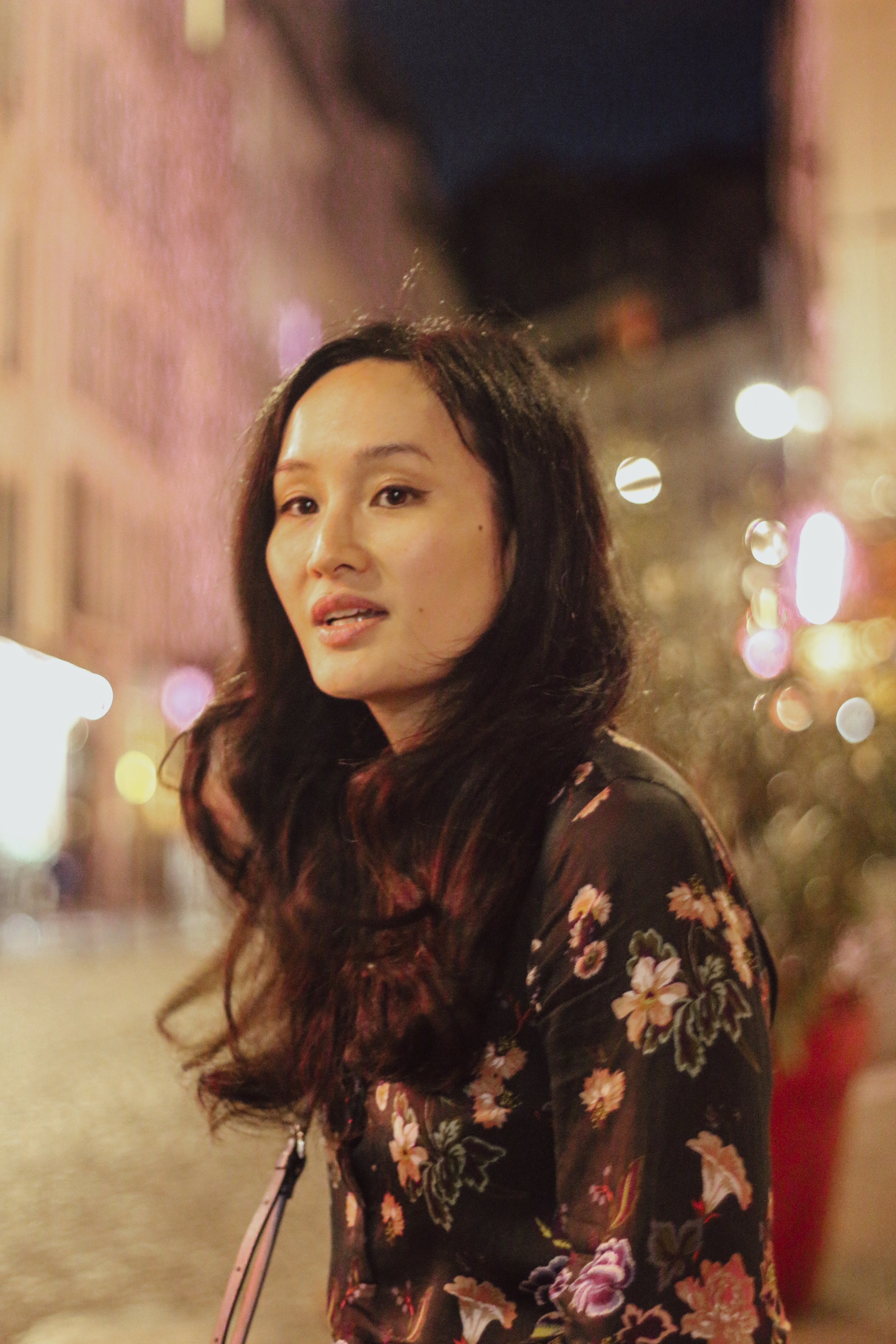 Une femme avec de longs cheveux noirs, portant une robe ou un manteau noir avec un motif floral, se tient dans une rue éclairée la nuit, avec un fond flou de lumières urbaines