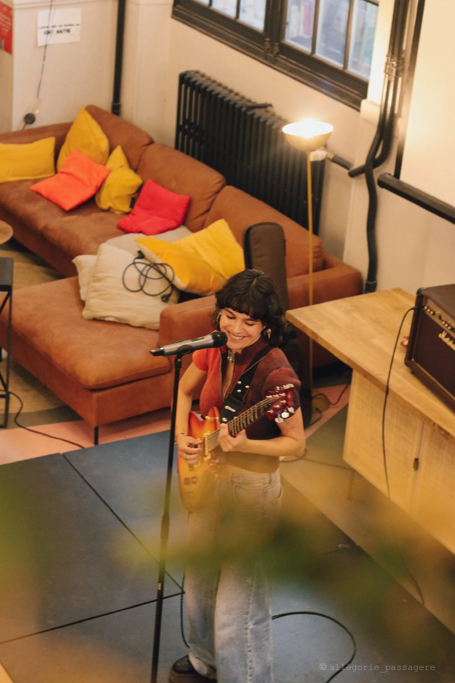 Une femme chante et joue de la guitare acoustique sur scène dans un espace intérieur, souriante, avec un microphone.