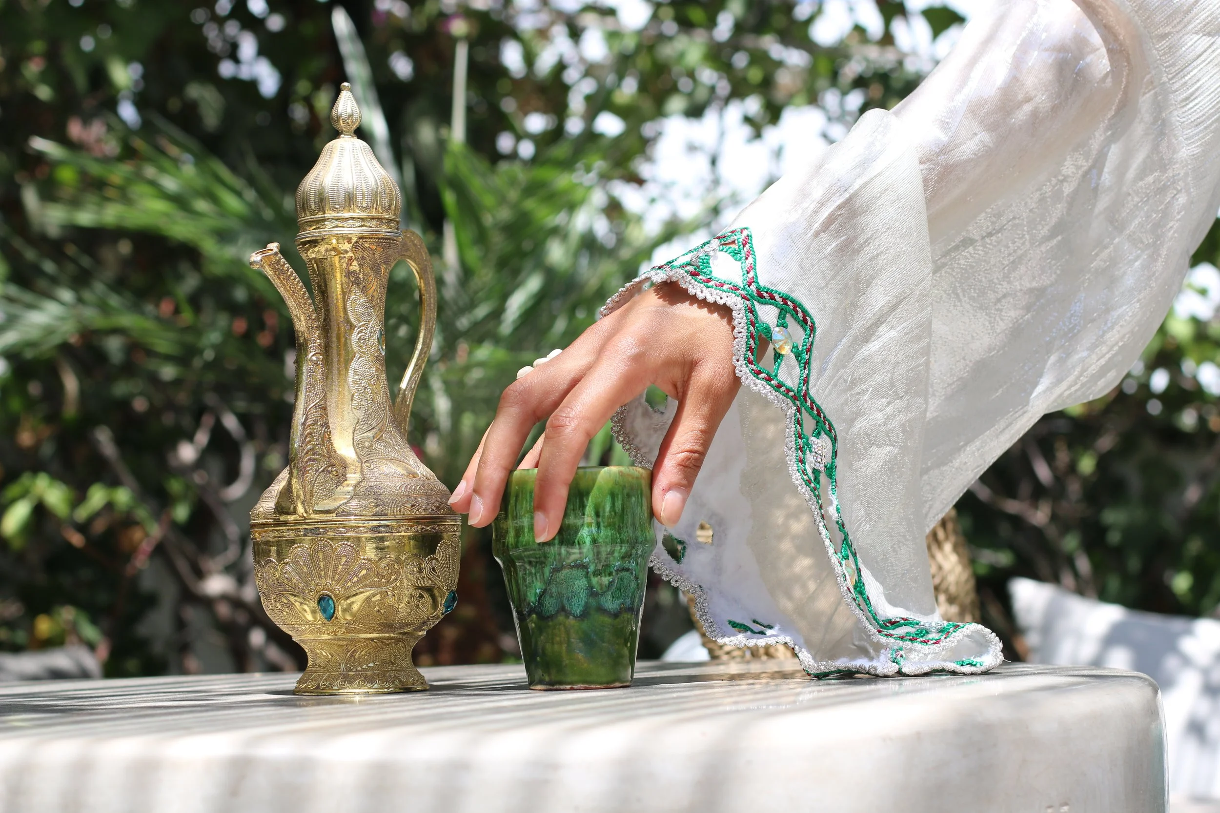 Une main en robe blanche manipule un verre en verre vert sur une table. À gauche, un vieux théière en métal doré avec des détails gravés et des pierres bleues.