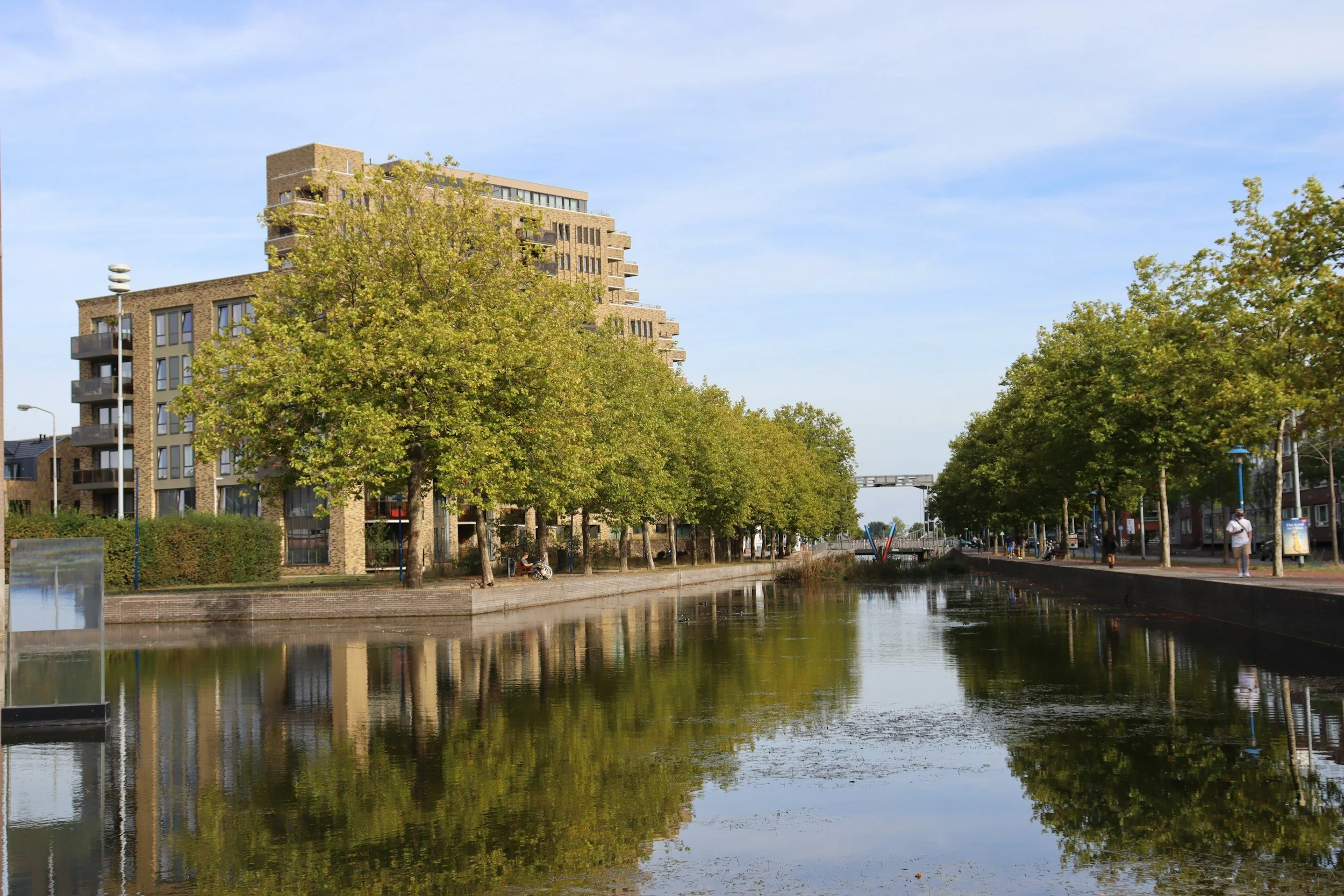 A cityscape with modern apartment buildings, trees lining a canal with reflections, pedestrians walking along the sidewalk, and a clear blue sky.