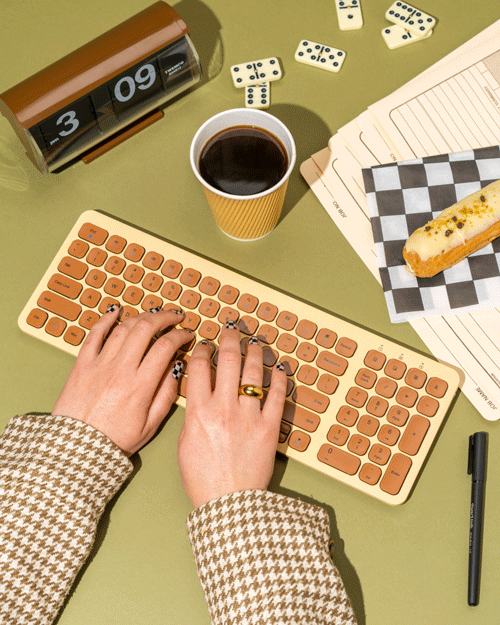 A person's hands typing on a peach-colored keyboard on a green desk. The desk also has a cup of coffee, a digital flip clock showing 3:09, some scattered dominoes, a notepad, a sandwich, and a black pen.
