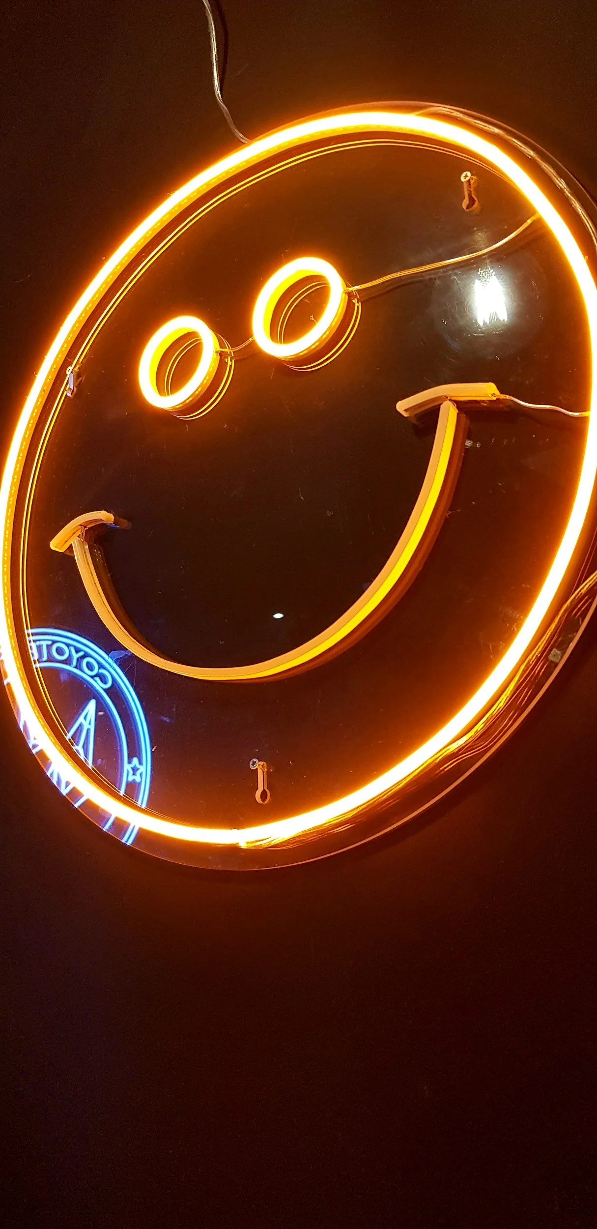 Neon sign of a smiling face with two round eyes and a curved mouth, glowing in yellow. A small blue neon circle with text is visible in the bottom left corner. The sign is mounted on a dark wall.