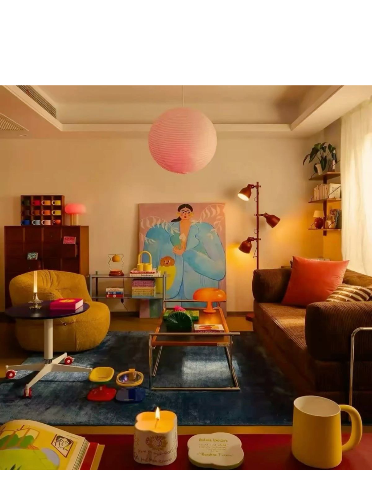 A cozy living room decorated with colorful furniture and playful decor. Features a large painting of a woman in traditional attire on the wall, a pink paper lantern hanging from the ceiling, a mustard-colored armchair, a brown sofa with pink and striped pillows, a dark blue rug, a coffee table with books and a lamp, children's toys scattered on the floor, a small side table with stacks of books, a tall floor lamp, and shelves with plants and decorative items.