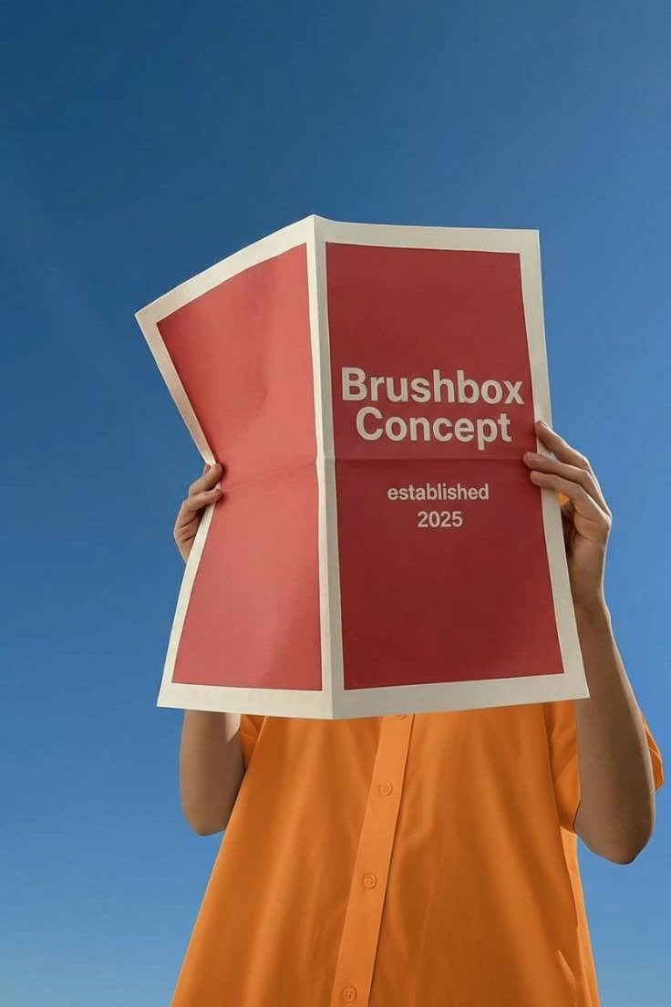 A person wearing an orange shirt holding a red and white brochure titled 'Brushbox Concept' against a clear blue sky.