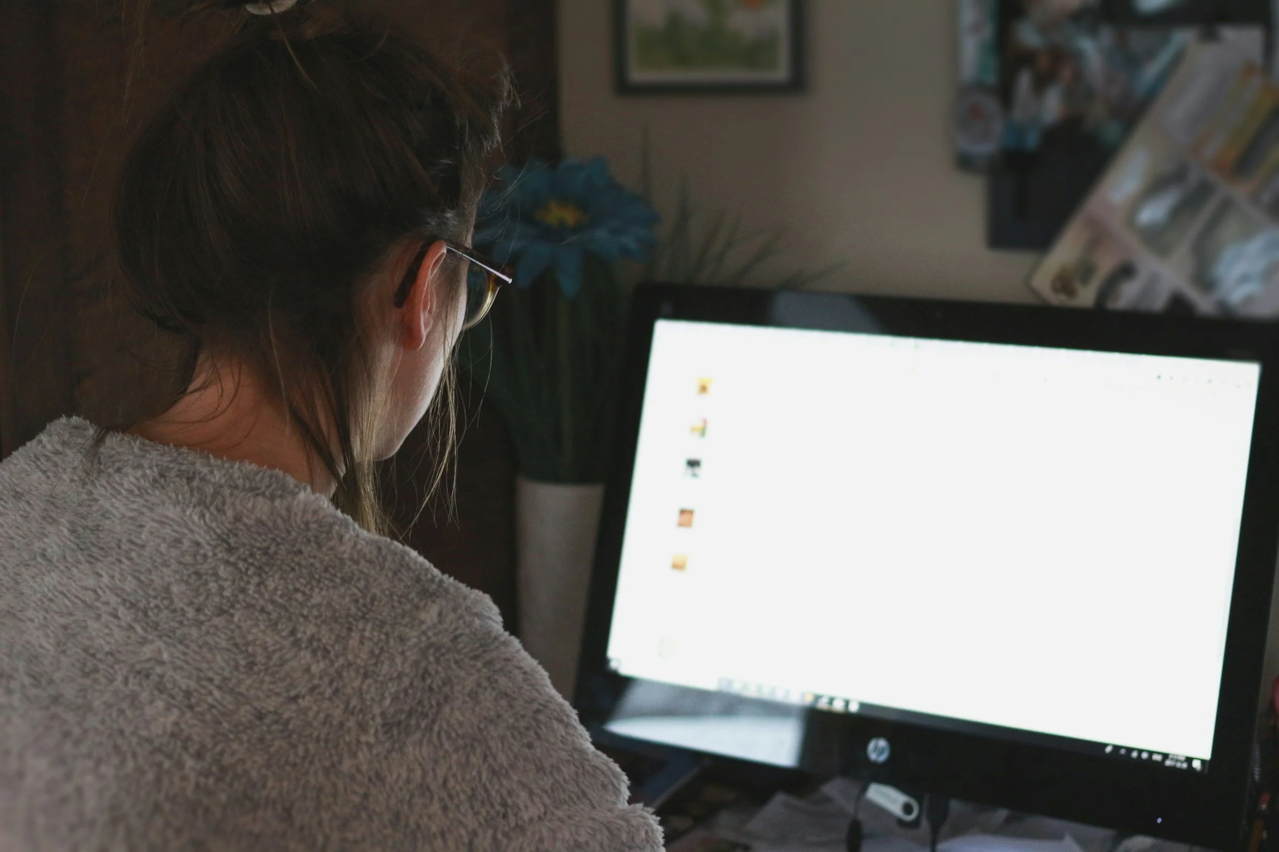 Person with brown hair wearing glasses and a grey fuzzy sweater working at a computer with a bright screen in a home office.