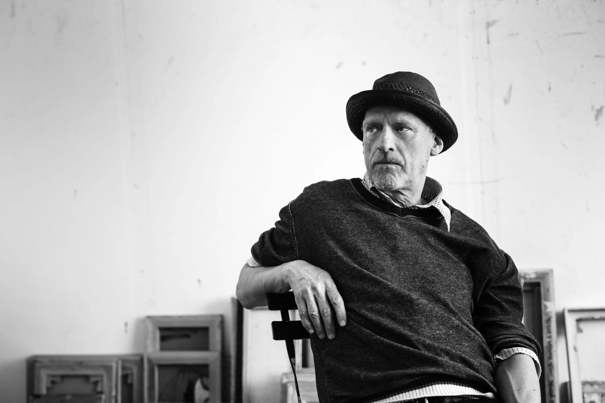 Sort-hvid foto af en mand, der sidder på en stol, iført sort hat og sweater, med en baggrund af tomme vægge og indrammede billeder eller tavler. Mandens ansigt er fokuseret og lidt op ad.