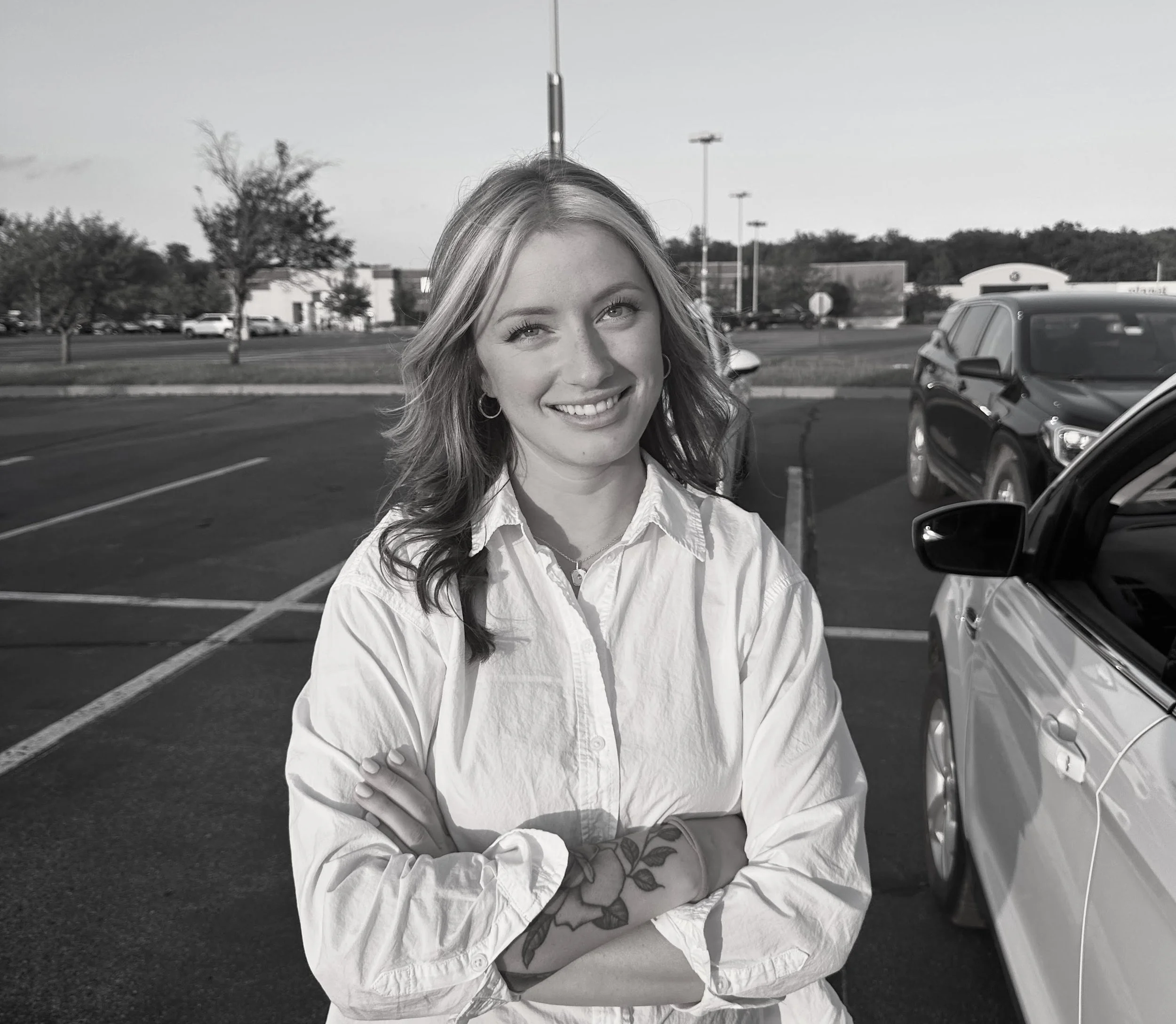 Black-and-white photo of a woman with wavy hair smiling, standing in a parking lot with cars and trees in the background.