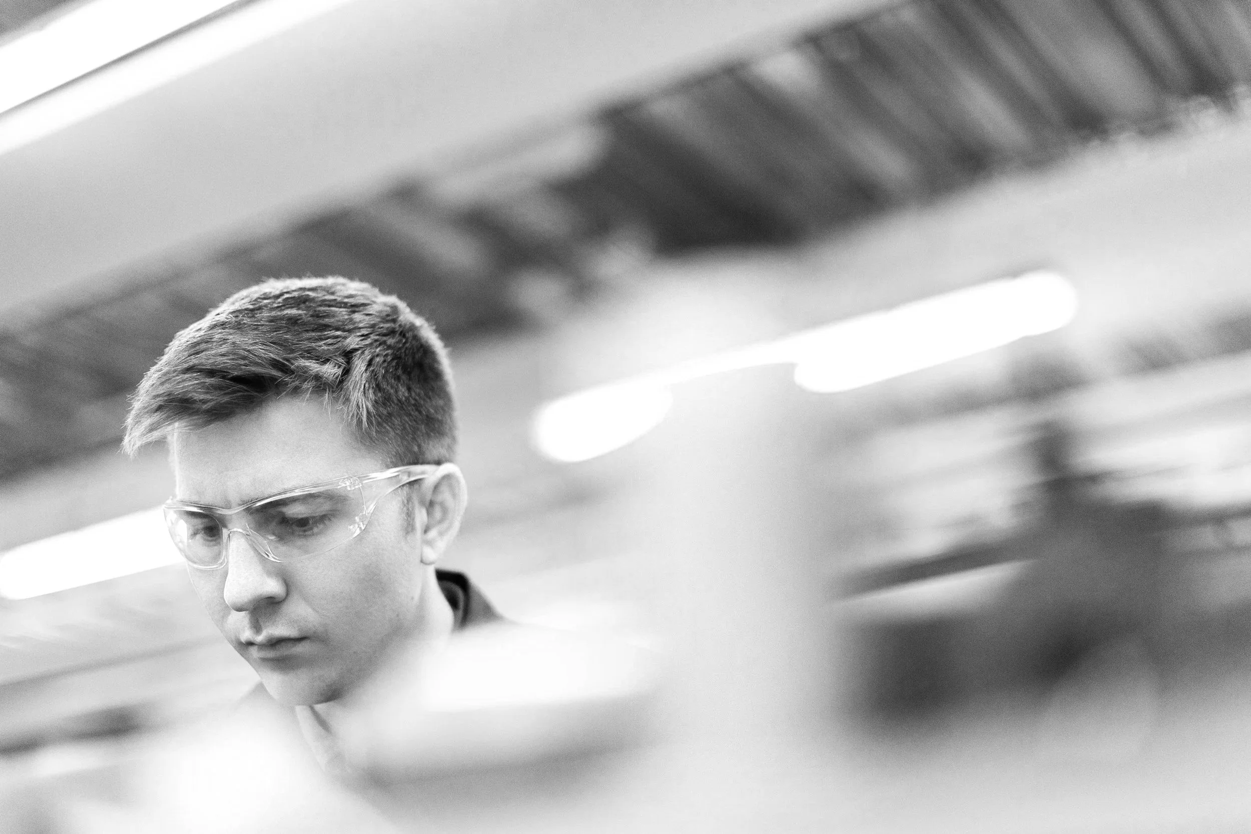 A young man with short hair and safety glasses working in a laboratory or industrial setting.
