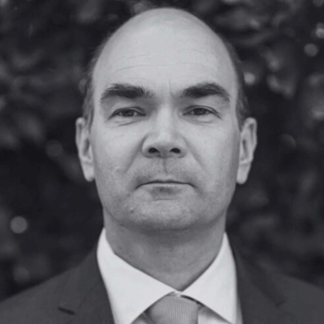 Black and white portrait of John Clarke wearing a suit and tie, with a serious expression, blurred background.
