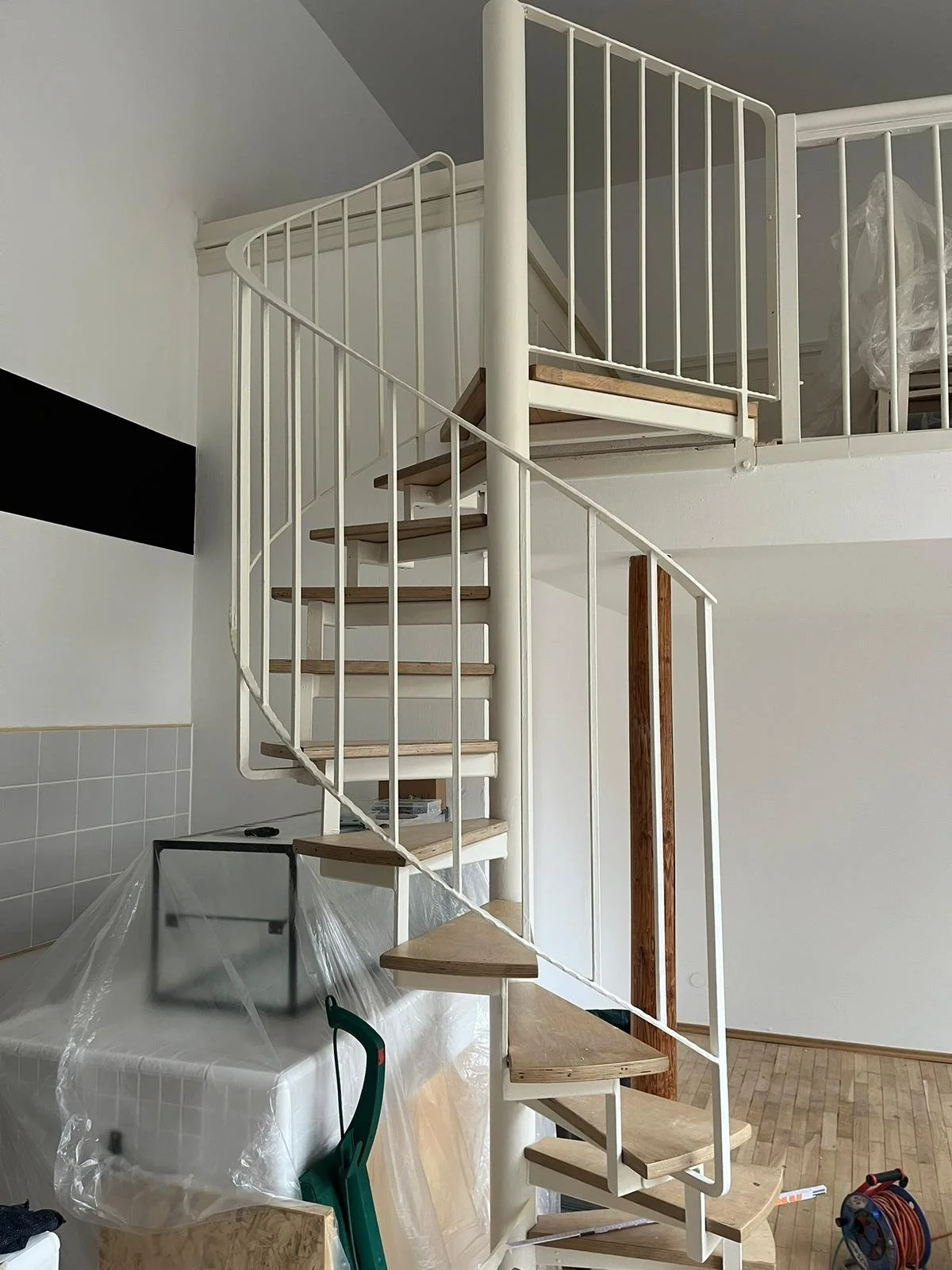 Innenaufnahme einer weißen Wendeltreppe mit Holzbodenstufen und weißen Geländern in einem modern eingerichteten Raum, der noch im Bau oder Renovierung ist.