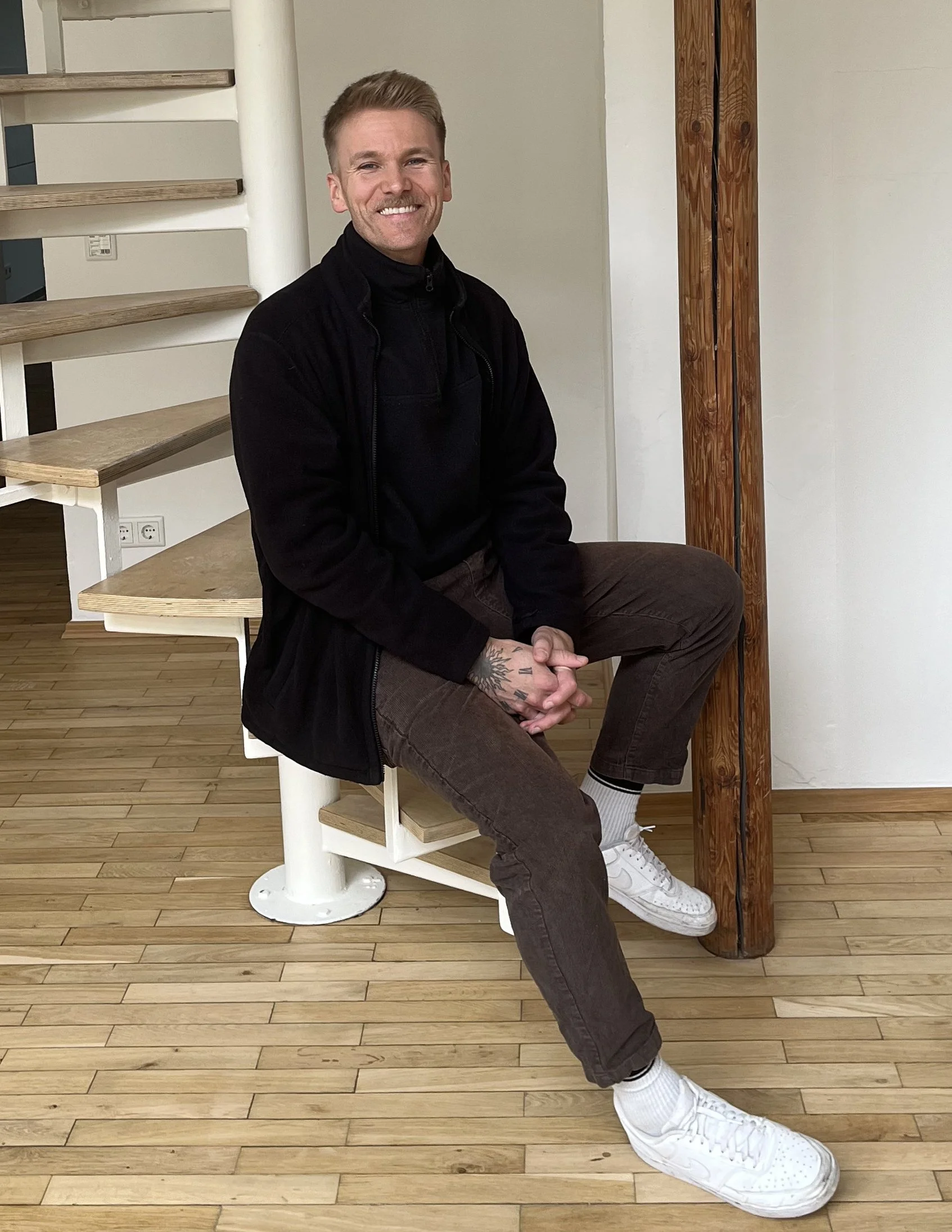 Ein junger Mann sitzt auf einer weißen Spindelstufe, lächelt und trägt schwarze Kleidung, weiße Sneakers und hat Tattoos am Handgelenk. Er befindet sich in einem Raum mit Holzboden und einer weißen Wand