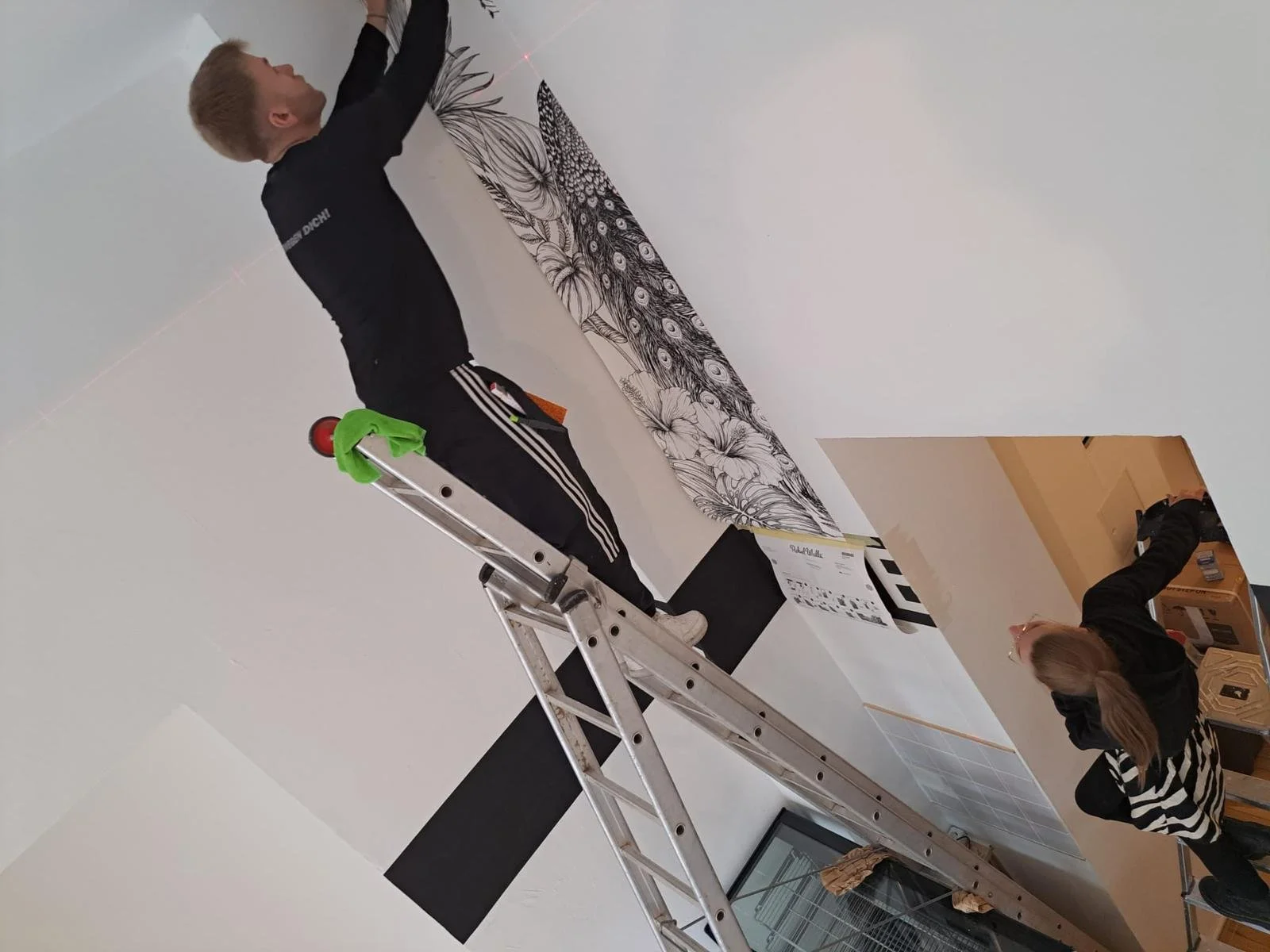 Zwei Menschen, eine junge Frau und ein junger Mann, hängen ein großes Wandbild mit Blumenmotiven auf einer weißen Wand auf, während die junge Frau auf einem Stuhl sitzt und die Frau auf dem Boden vor einem Karton arbeitet.