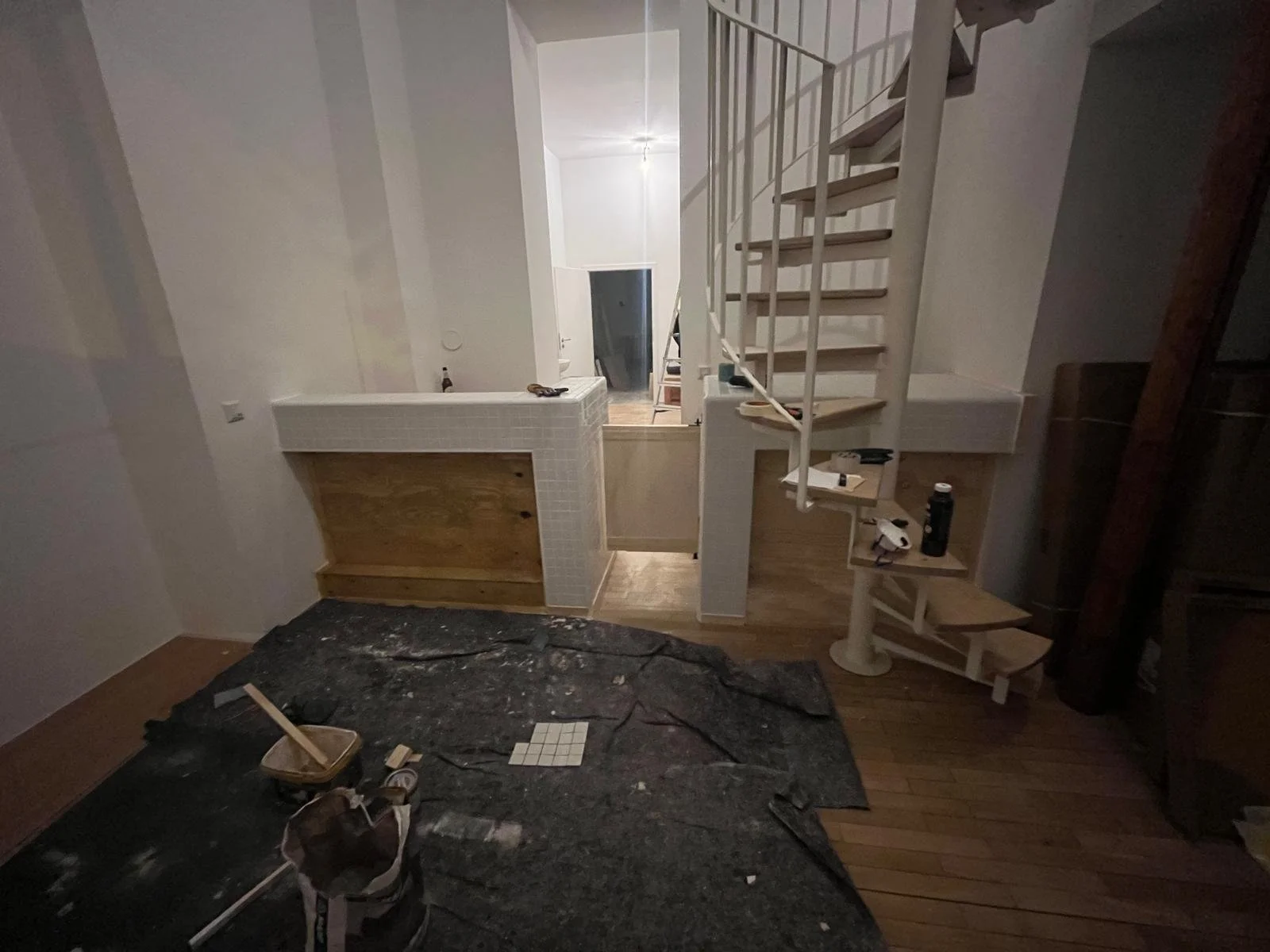 Innenaufnahme eines Hauses unter Renovierung, mit einer Wendeltreppe aus Holz und einer halb aufgebauten Bar oder Theke, auf dem Boden liegt eine Abdeckung mit Werkzeugen und Malerutensilien.