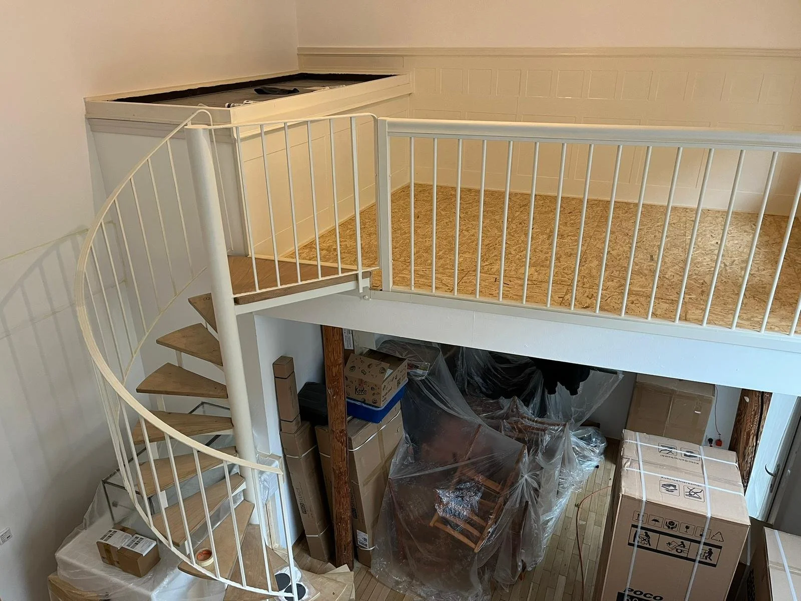 Innenraum mit weißer Treppe, die nach oben führt, und einem offenen Raum darunter mit Kartons und Verpackungsmaterial.