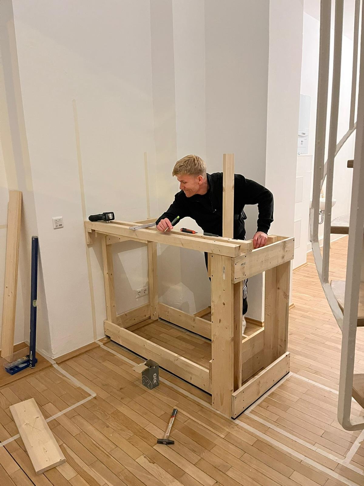 Ein Mann arbeitet an einem Holzrahmen, vermutlich beim Möbelbau, in einem Raum mit Holzboden.