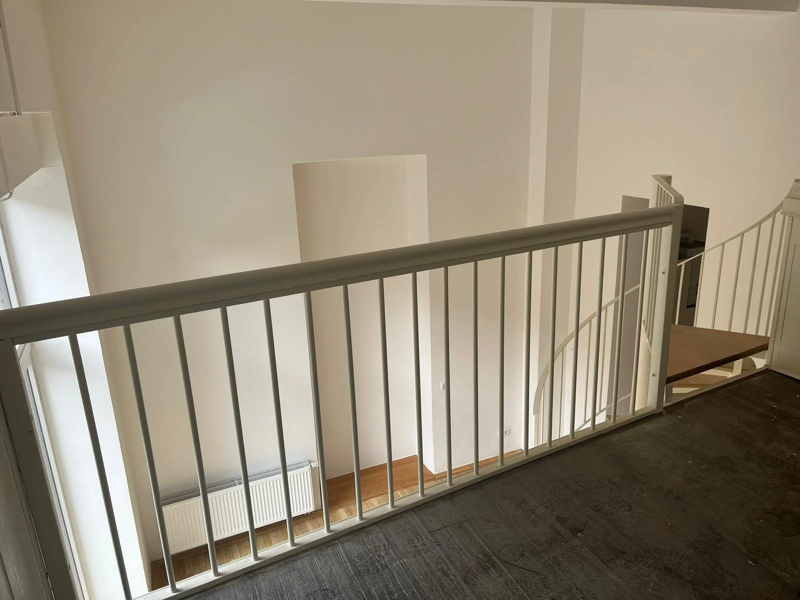 Eine weiße Innentreppe mit Geländer führt nach unten in ein hell erleuchtetes Zimmer. Das Bild ist von oben aufgenommen, zeigt den oberen Balkon und die Wand mit einem Heizkörper.