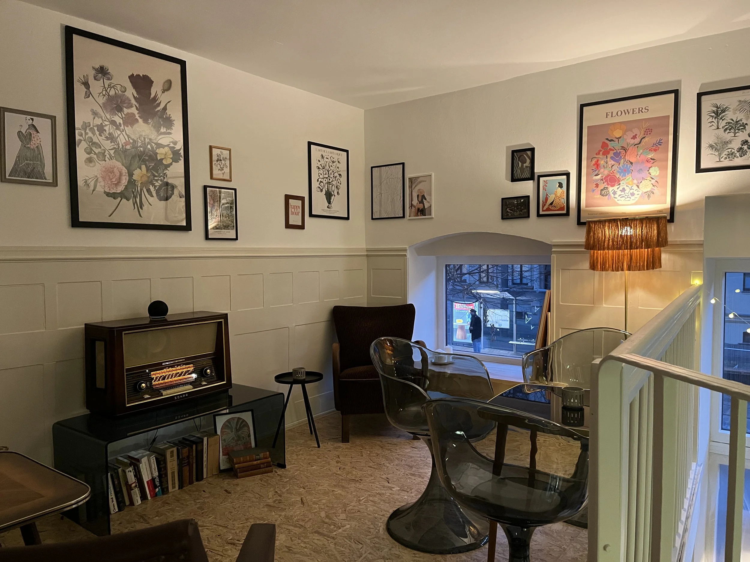 Gemütliches Wohnzimmer mit Wandbildern, Sofas, Couchtisch, Sessel, einem Fenster mit Blick nach draußen und einer Stehlampe mit braunem Lampenschirm.